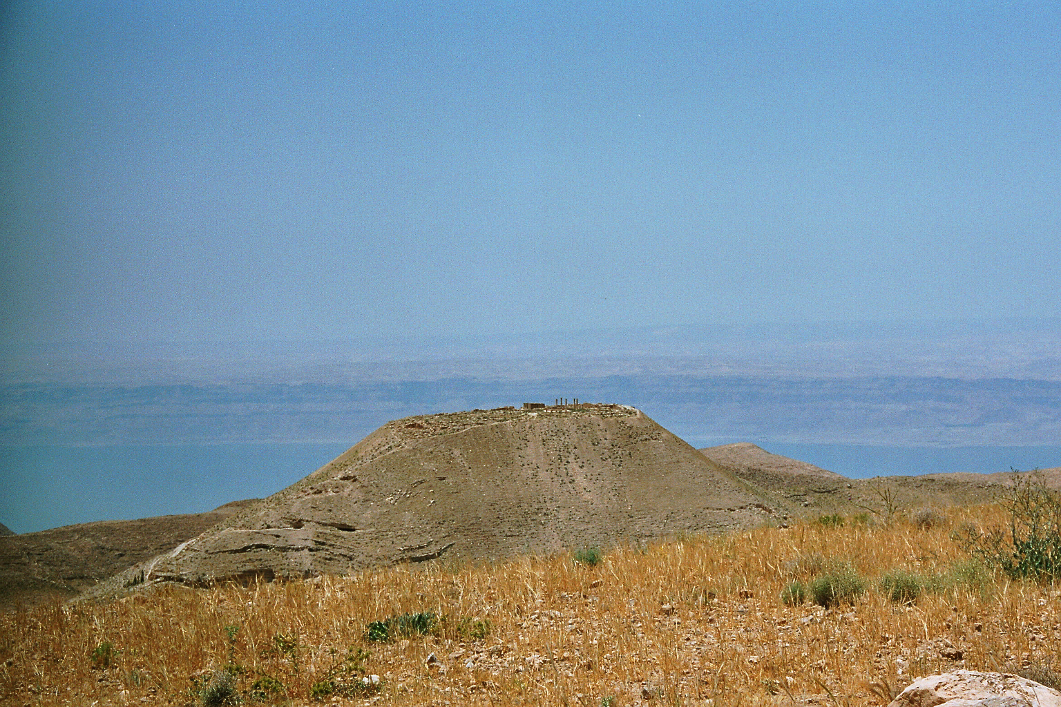 Panoramic photograph of a bare hilltop with the low stone remains of a fortress, surrounded by dry, steep hills