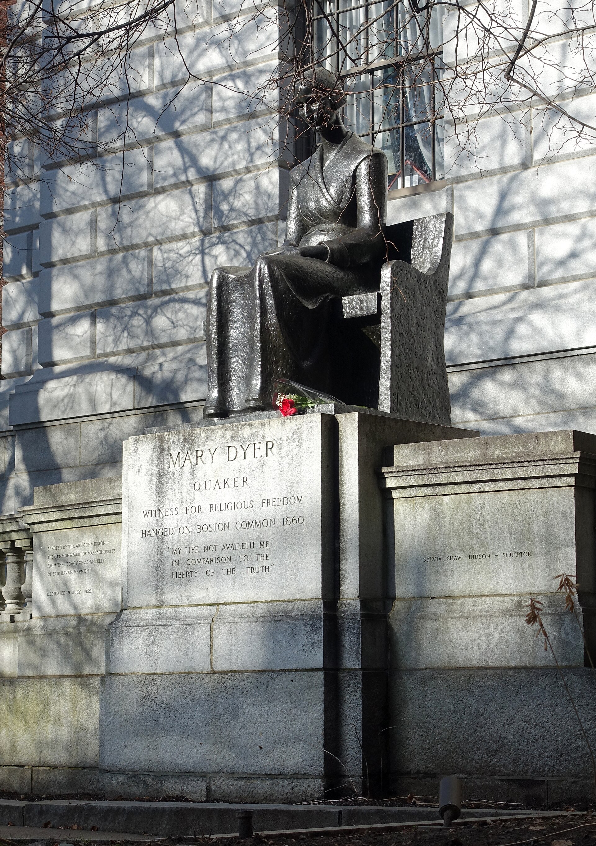 A bronze statue of a seated woman in a plain bonnet and long dress, hands folded in her lap, on a stone base in front of a granite building.