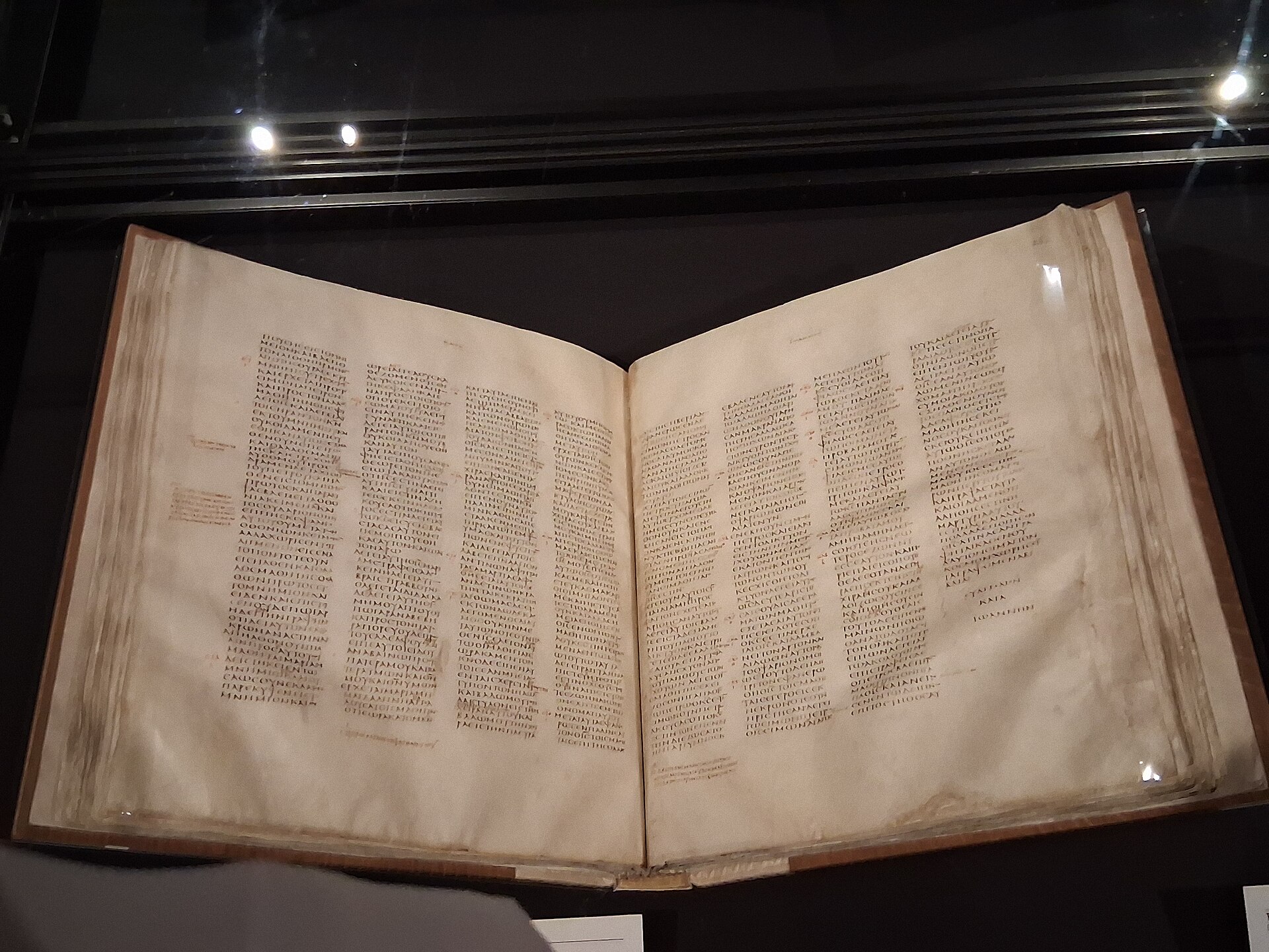 An open ancient manuscript with pages of Greek capital letters in narrow columns, displayed in a glass case in a dimly lit gallery
