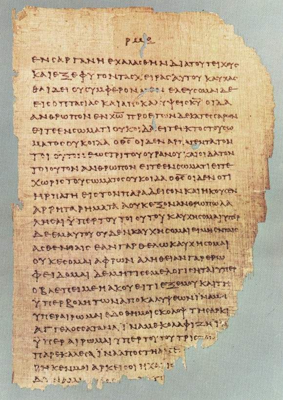 A tall, worn papyrus page with a single column of Greek capital letters, its edges broken and its lower part damaged