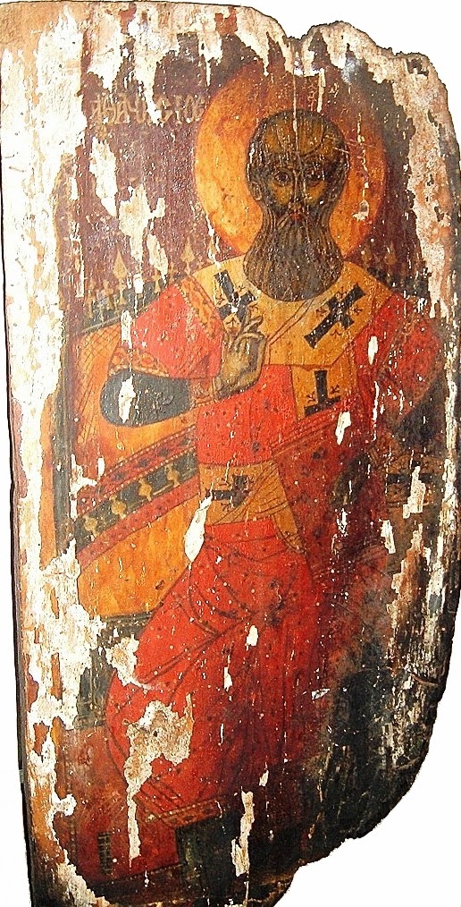 A tall painted icon of a bearded bishop in patterned vestments with a halo, holding a closed book and raising his right hand in blessing