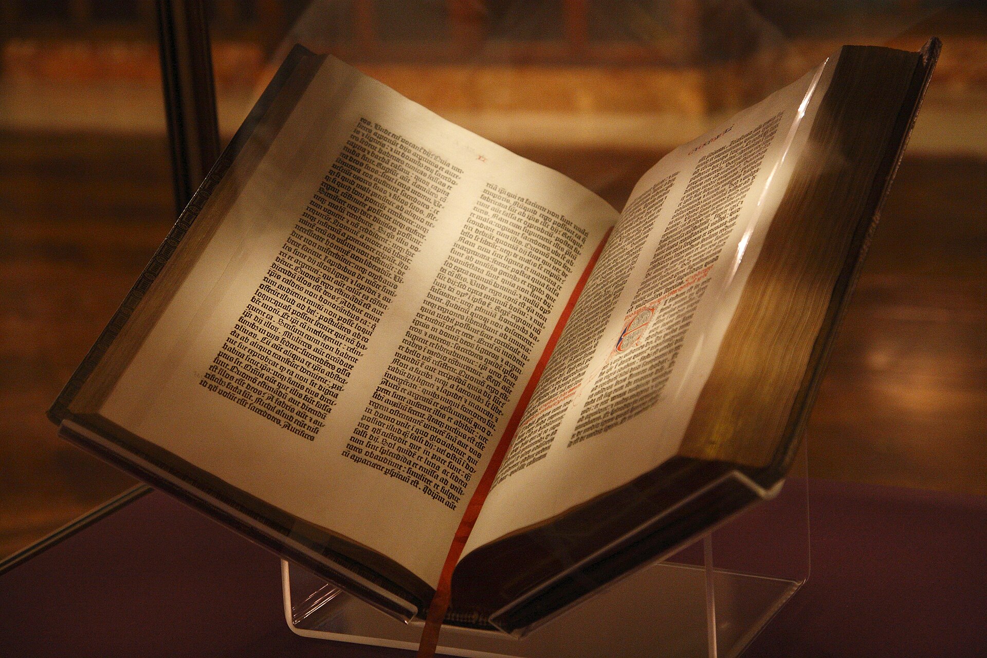 An open printed Bible in a glass display case, its two pages set in two dense columns of black Gothic type with large colored initials