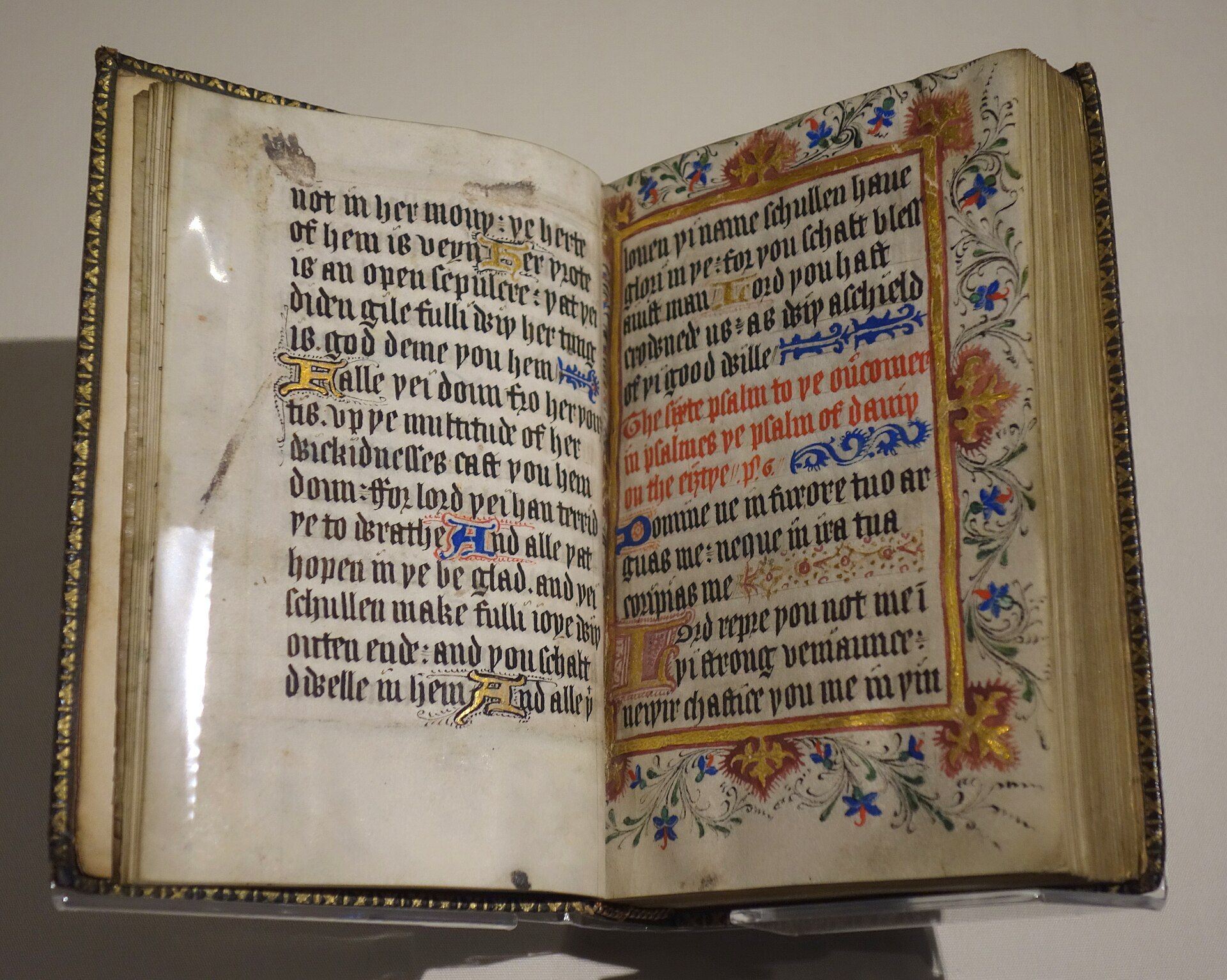A small open manuscript book in a museum case, showing two pages of brown handwriting with red and blue decorated capital letters
