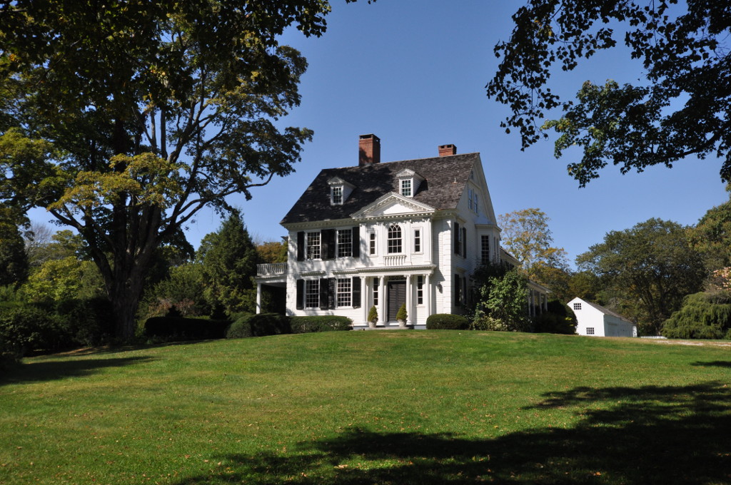 A two-story white clapboard colonial house with a central chimney and many-paned windows, set behind a lawn and trees