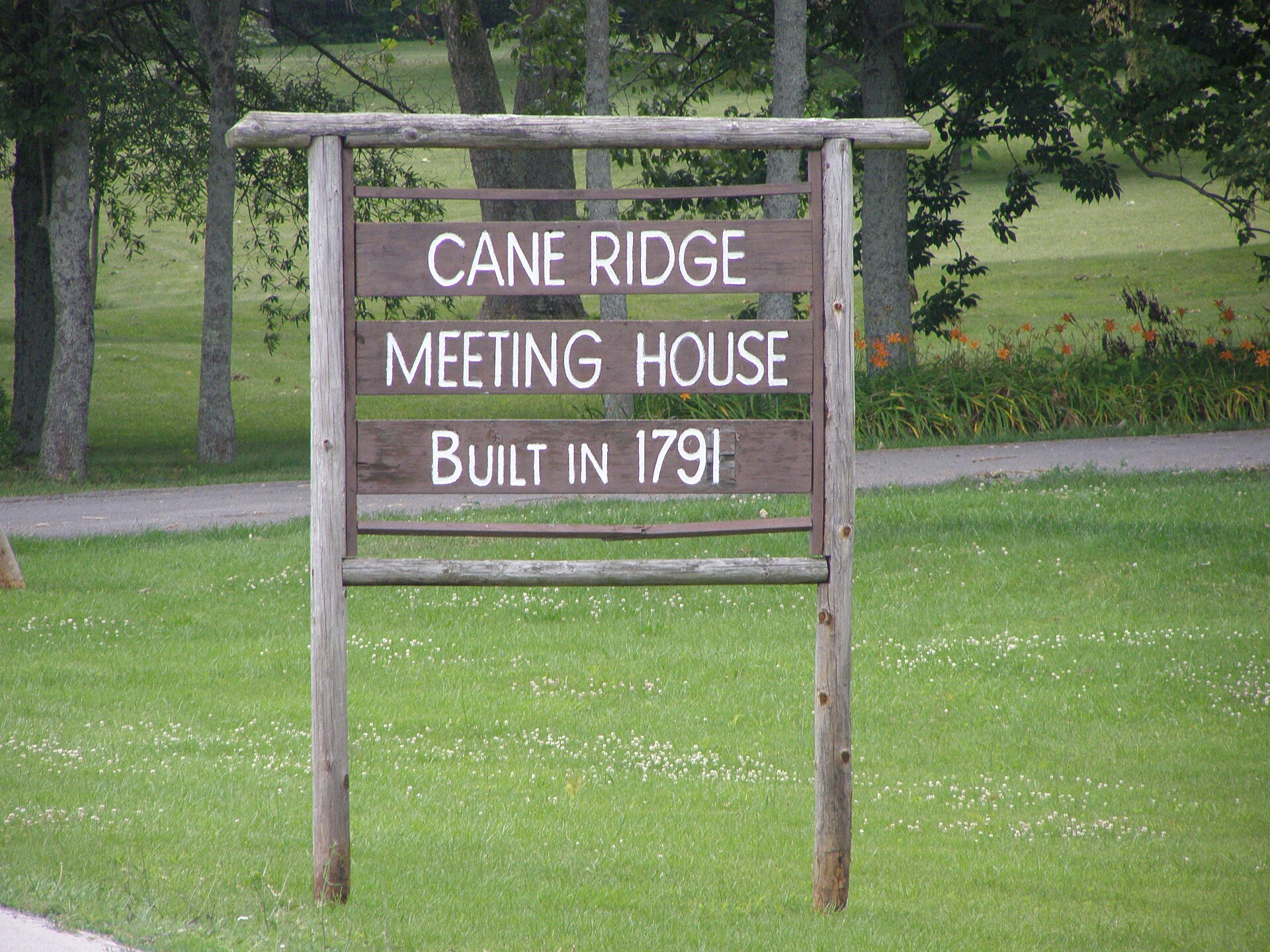 A two-story log building with a wood shingle roof and small windows standing inside a large modern stone and glass enclosure