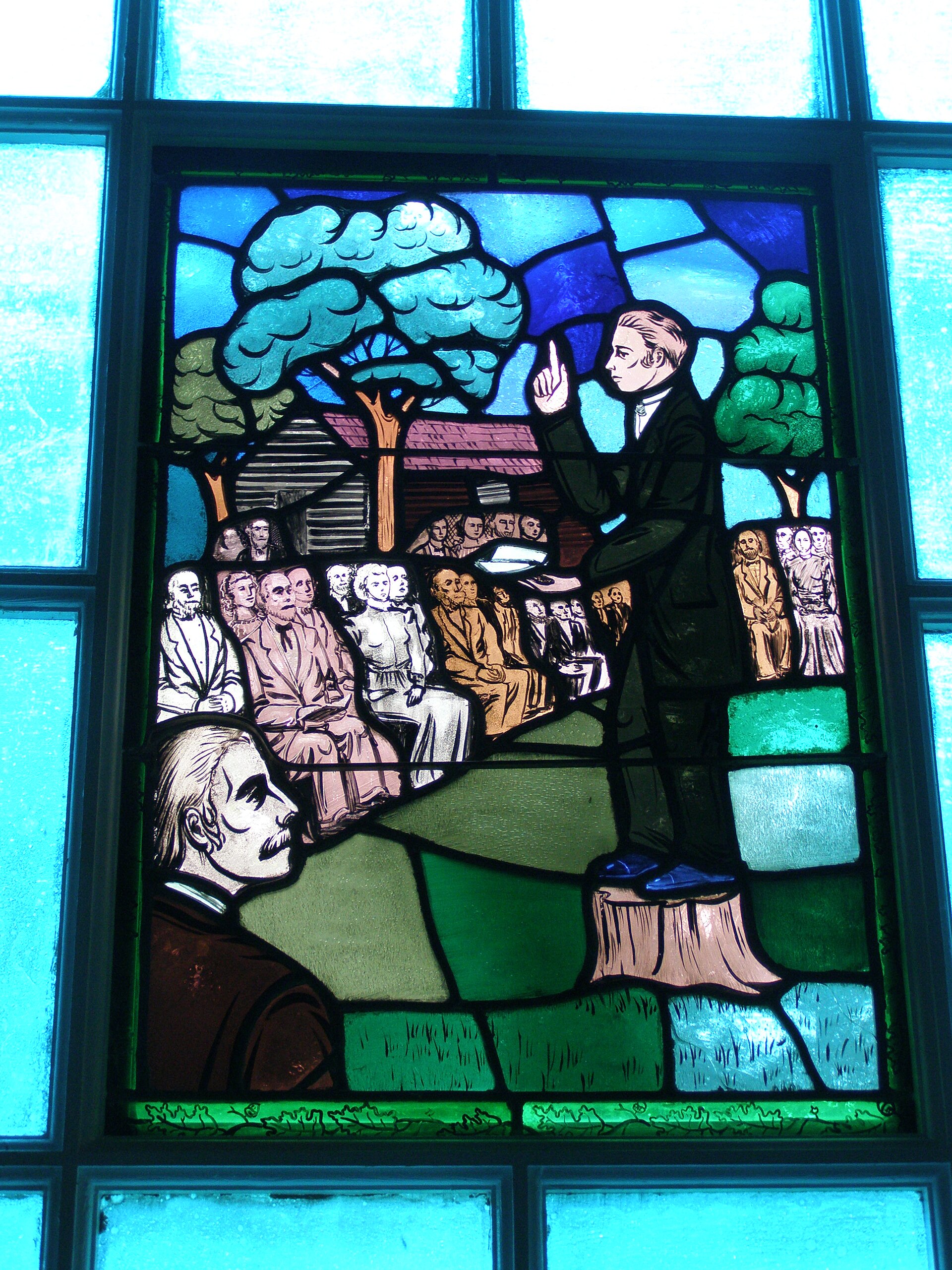A round bronze-colored medallion in relief showing a preacher standing on a stump with raised arm before a seated and standing crowd among trees