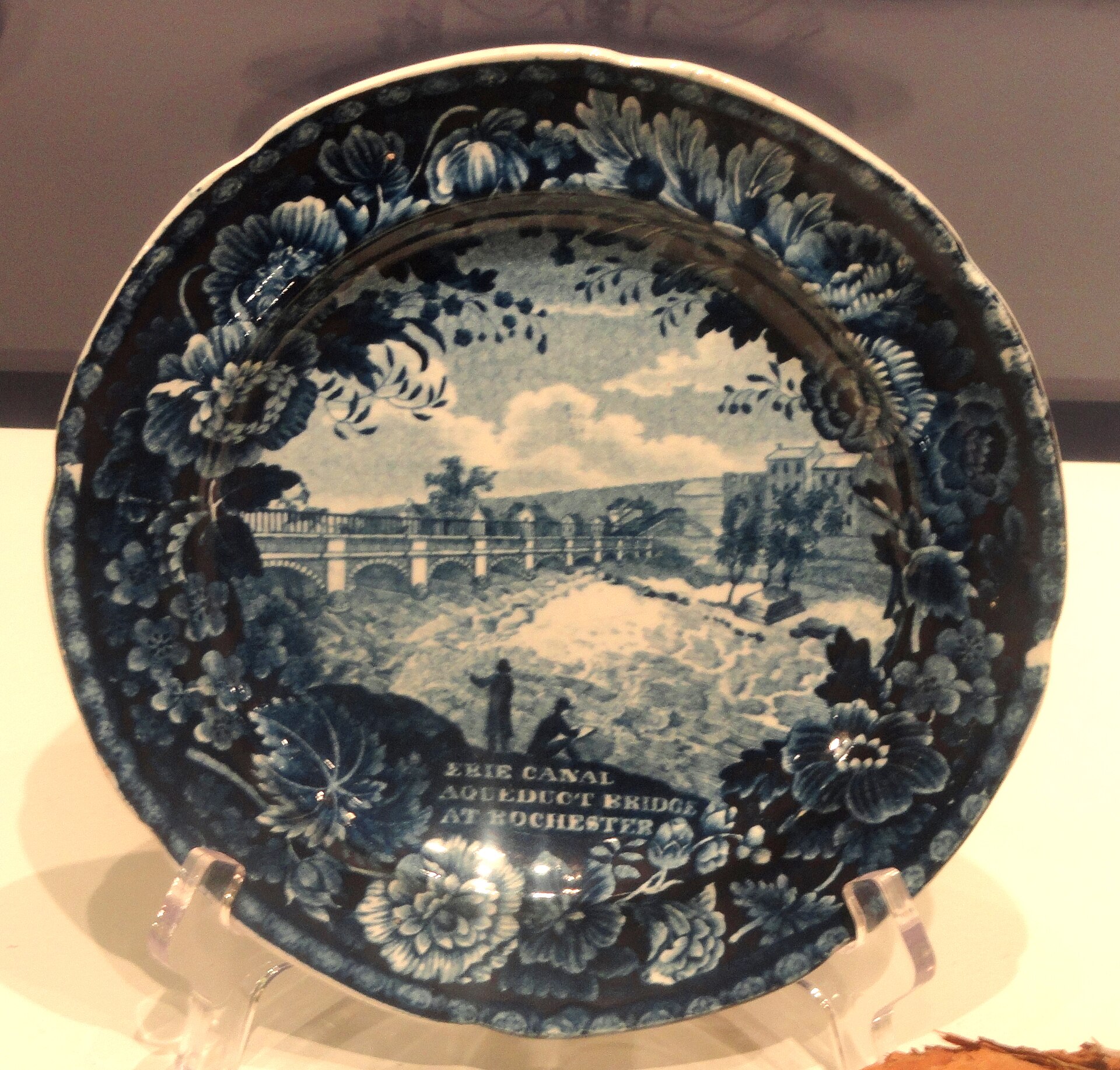 A blue and white transfer-printed ceramic plate with a scene of a canal, boats and buildings in the center and a border of flowers and shells