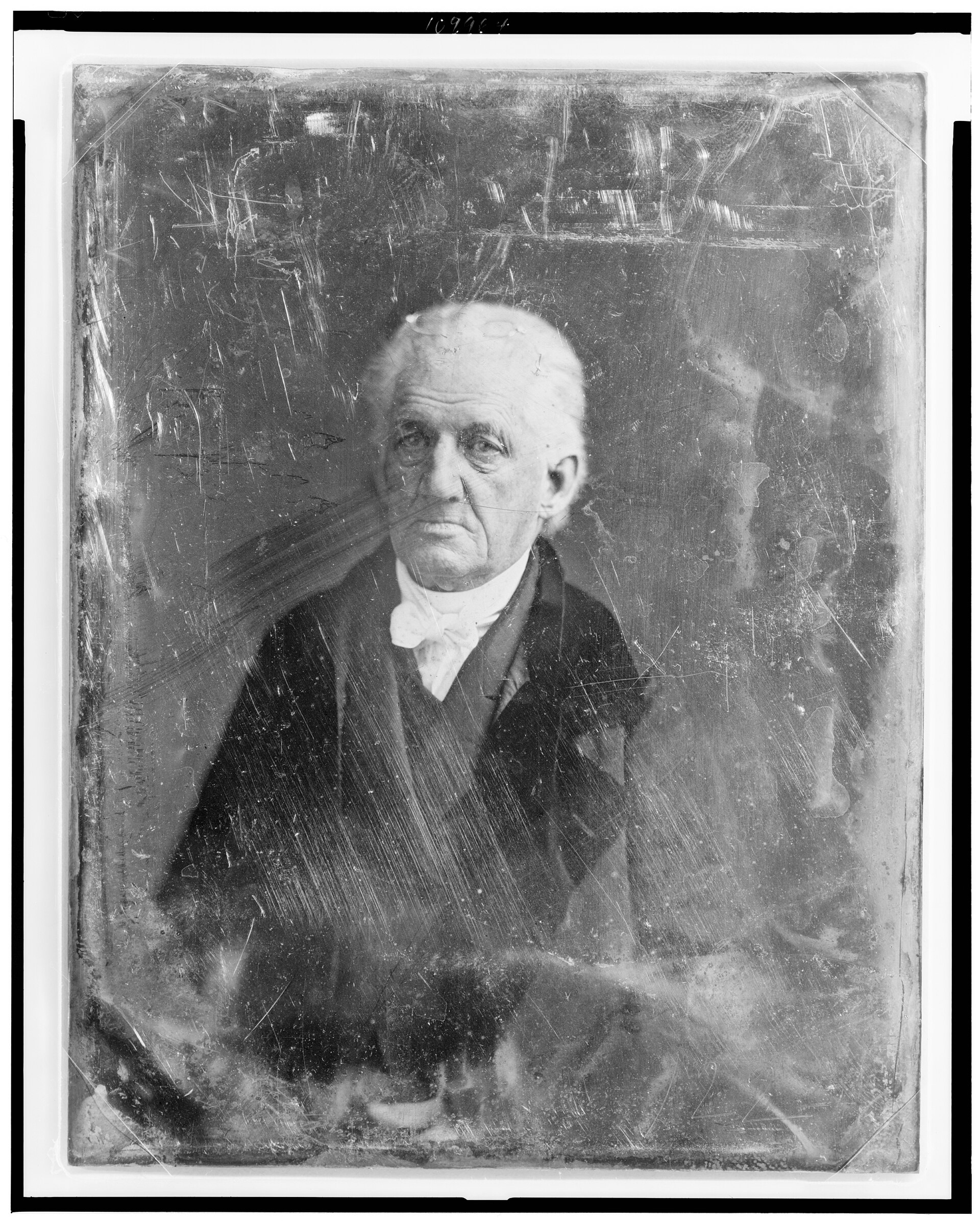 Photograph of an elderly man with swept back gray hair, seated, in a dark coat with a high collar, looking directly at the camera