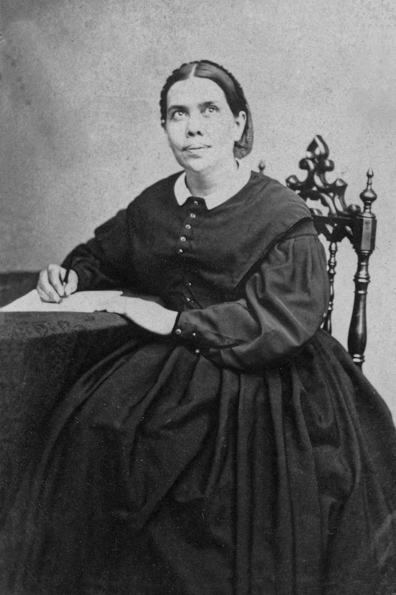 Photograph of a woman in a dark dress seated in an ornate chair at a small table, holding a pen over paper