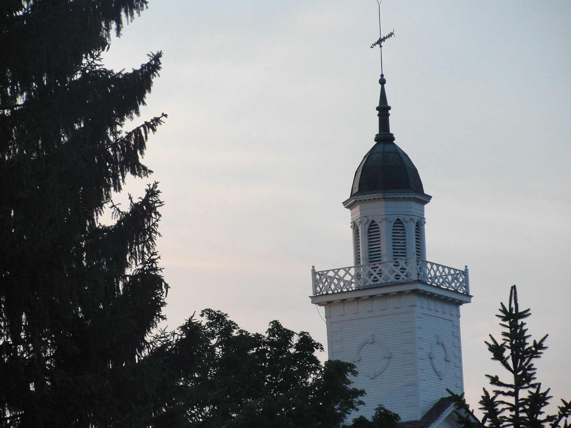 A two-story white stone and stucco church building with tall arched windows and a square tower with a steeple, on a lawn under a blue sky