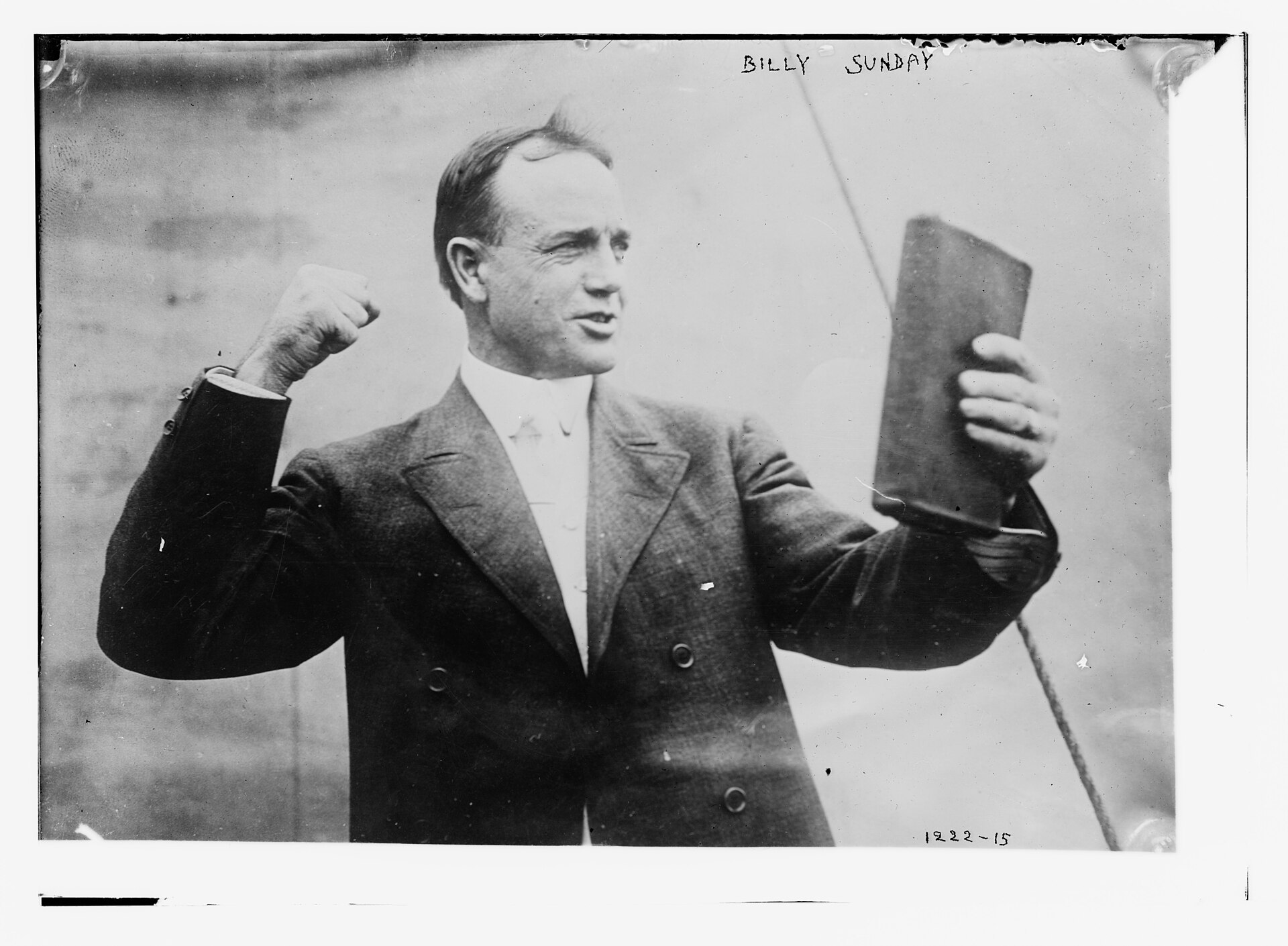 Black and white photograph of a man in a dark suit raising one fist and holding up a book in the other hand, as if preaching