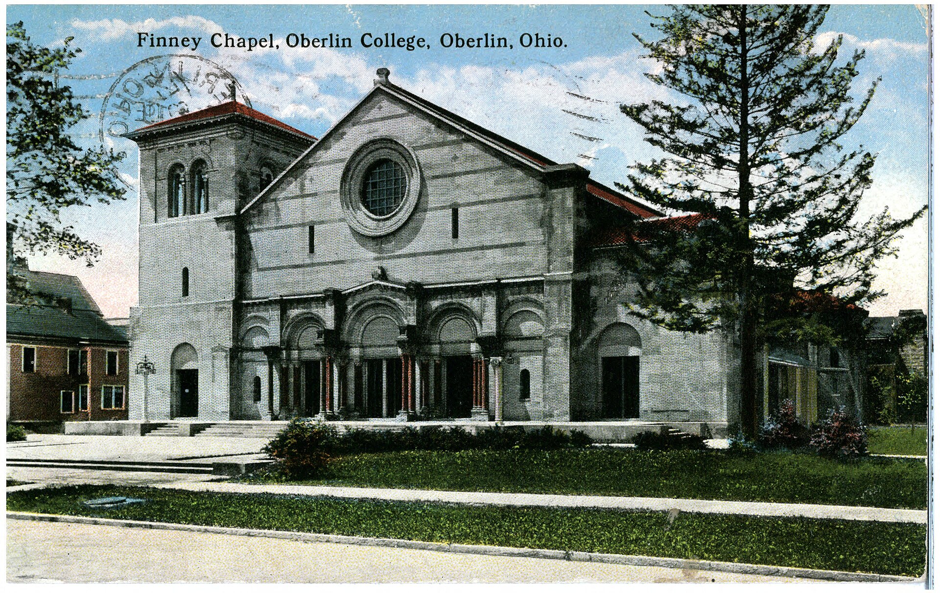 Colored postcard view of a large stone church with a round window in its front gable, a row of arched doorways and a square tower at the left, with a lawn and a tall evergreen tree in front