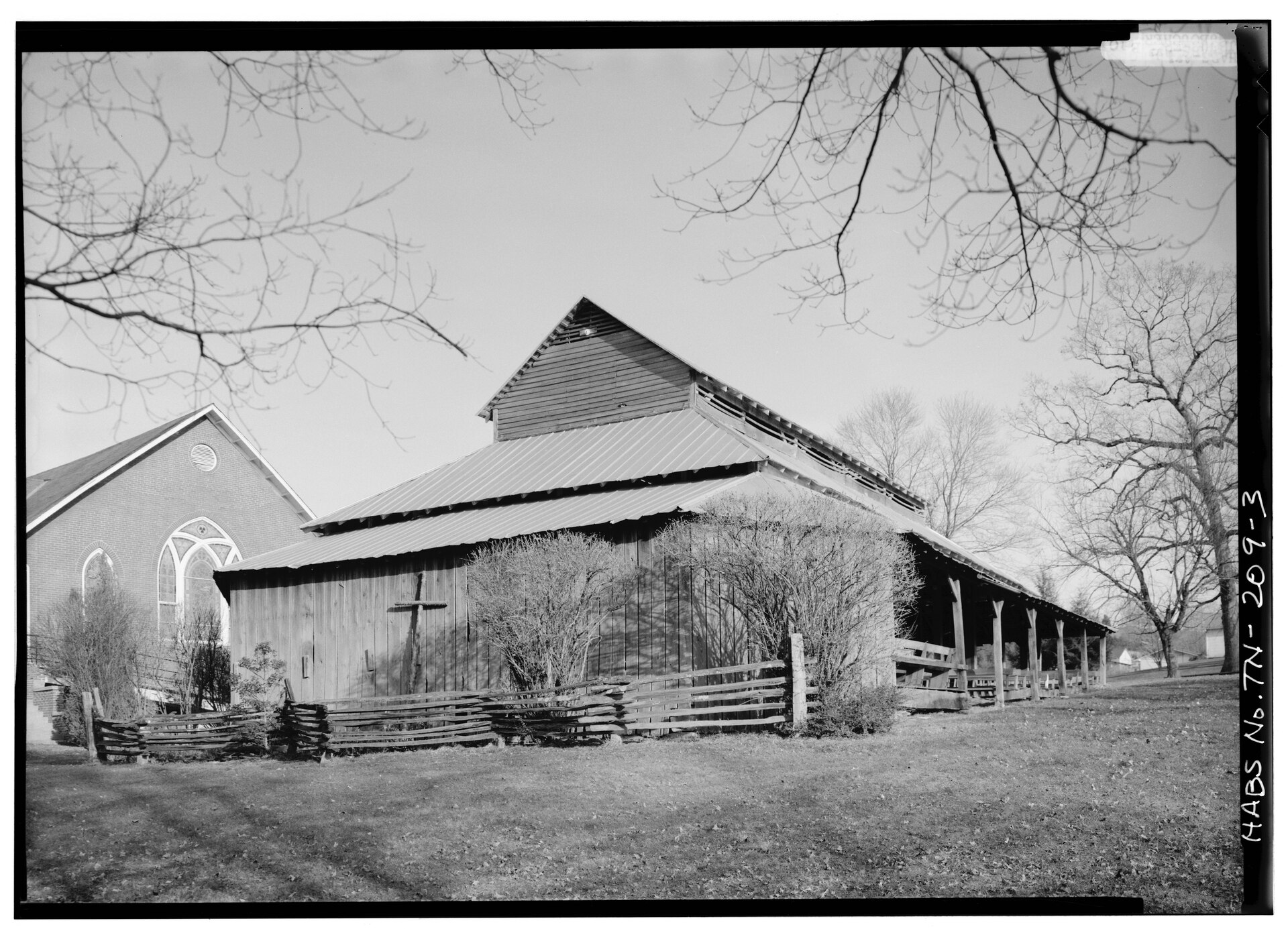 Black and white photograph of a long wooden building with a raised roof and a wooden cross on its side wall, behind a split-rail fence, with a brick church with arched windows at the left and bare trees around