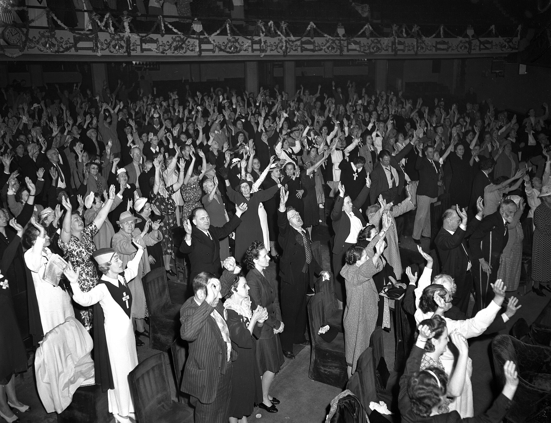 Black and white photograph of a large domed auditorium filled with worshipers on the main floor and in curved balconies, many with raised hands, facing a platform
