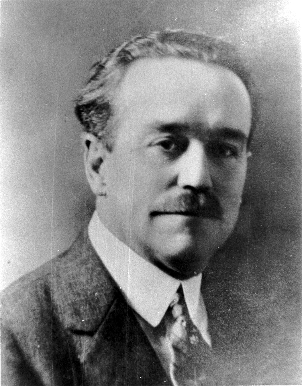 Black and white head and shoulders portrait of a man with a mustache and short dark hair, wearing a suit jacket, white collar and tie