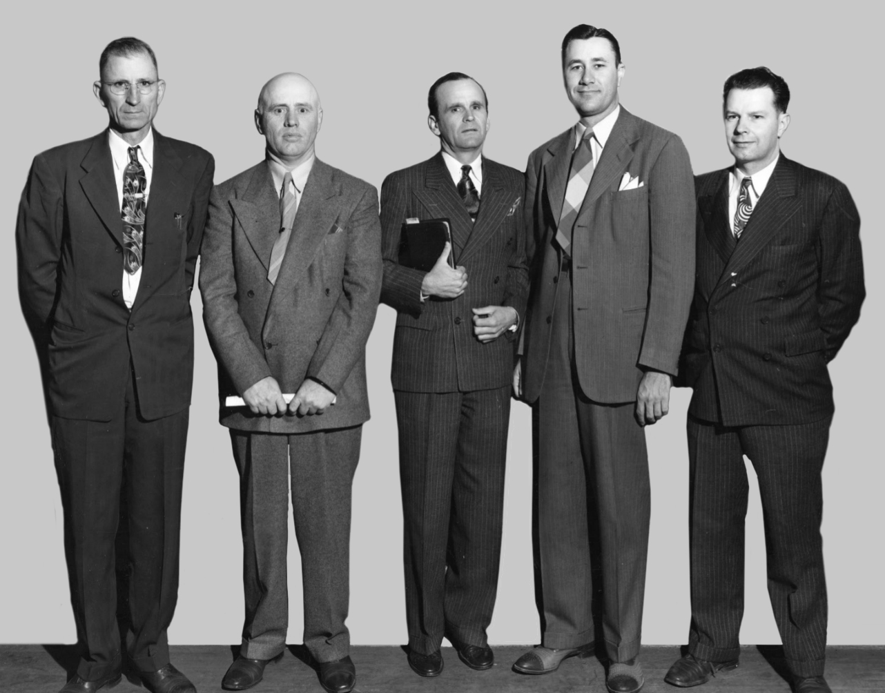 Black and white photograph of five men in suits and ties standing side by side indoors