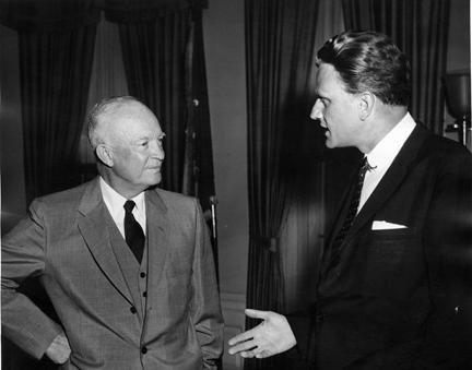 Black and white photograph of President Dwight Eisenhower, in a light suit, talking with Billy Graham, in a dark suit, in front of dark curtains