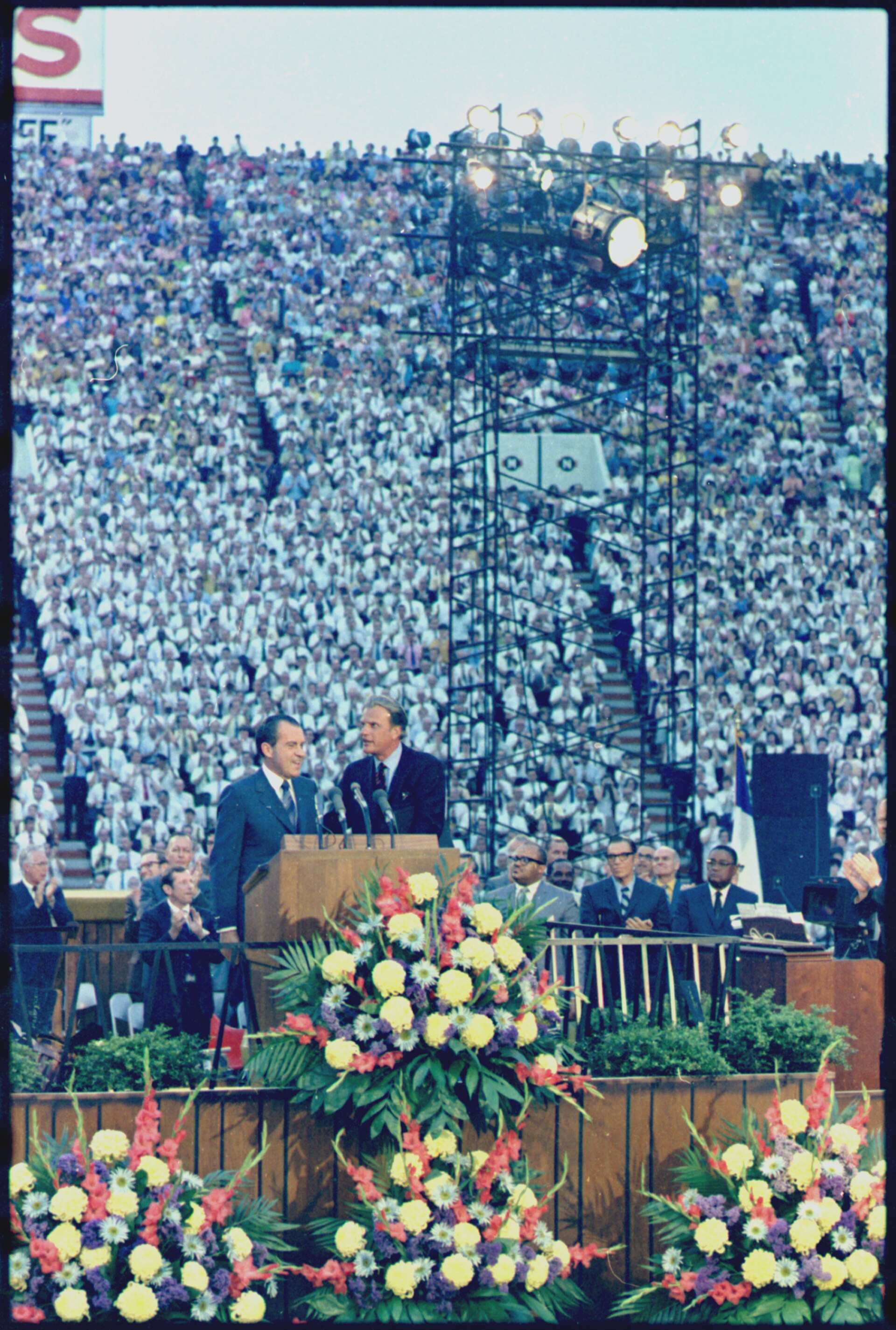 Color photograph of a packed stadium; at a lectern decorated with banks of flowers, Richard Nixon stands beside Billy Graham on a platform