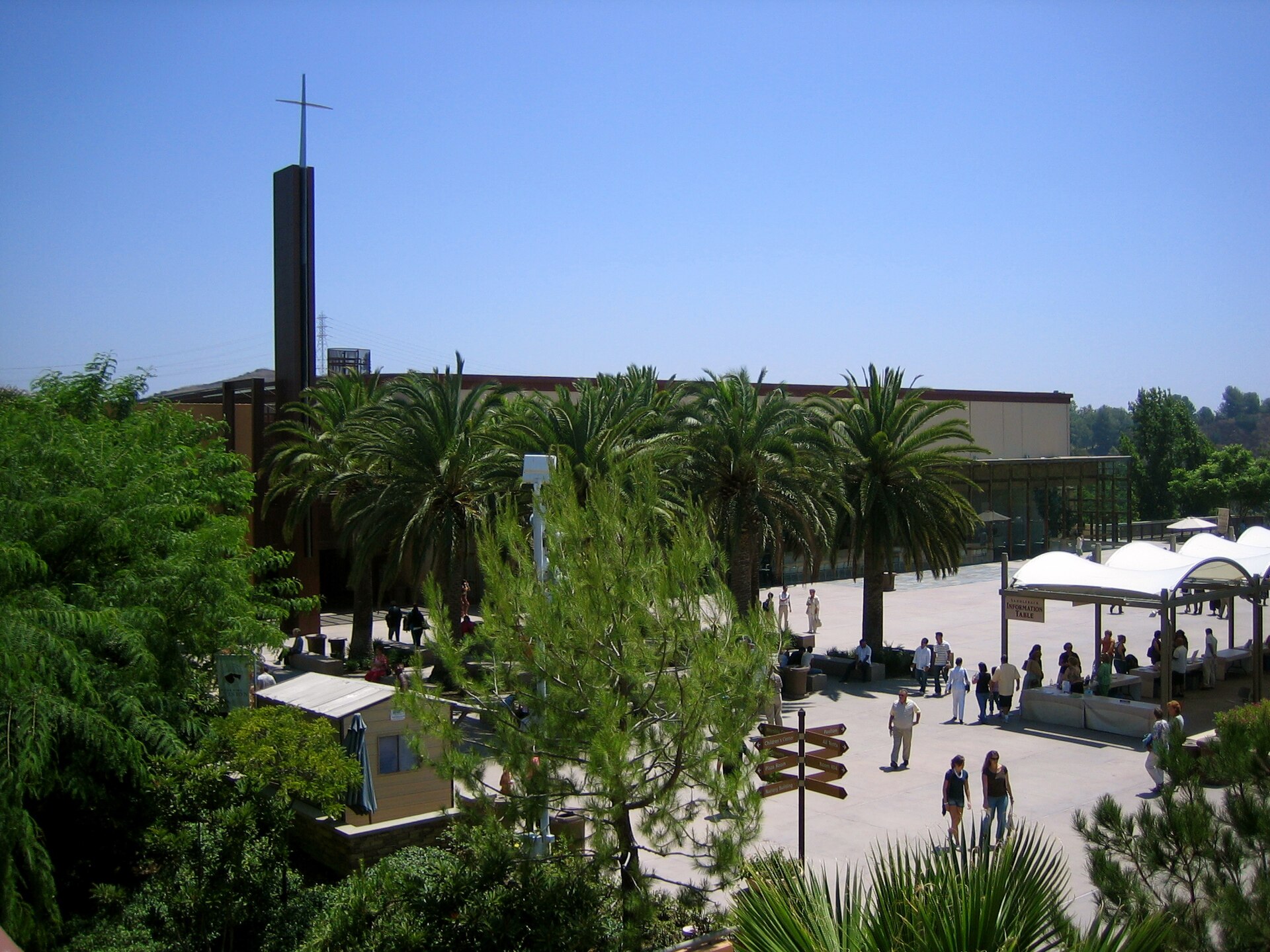 An elevated view over palm trees to a paved plaza with people and white canopies, beside a large low building with a tall tower topped by a cross