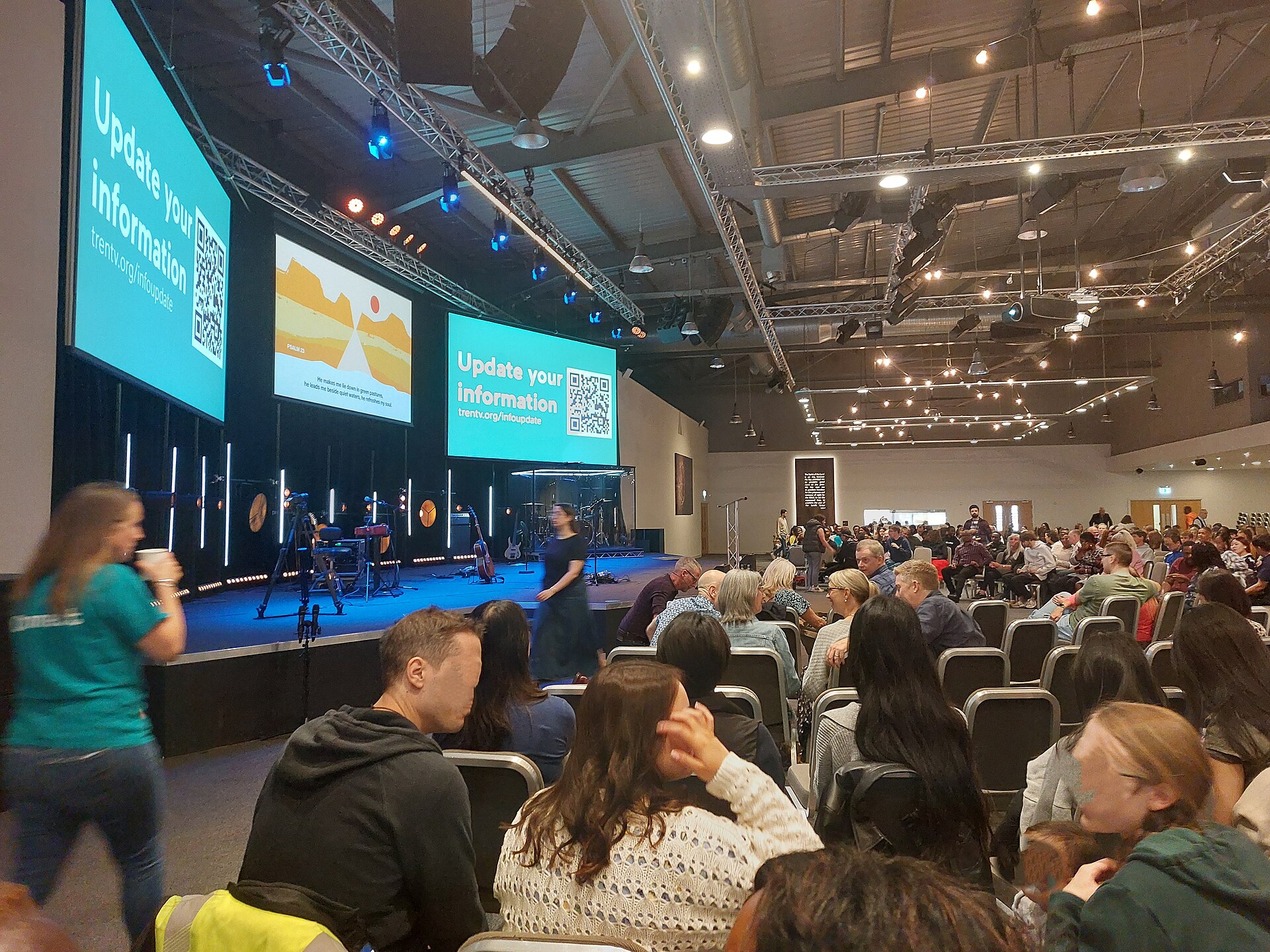 People stand and talk inside Trent Vineyard during a break in a Sunday service; the faces of those nearest the camera are blurred.