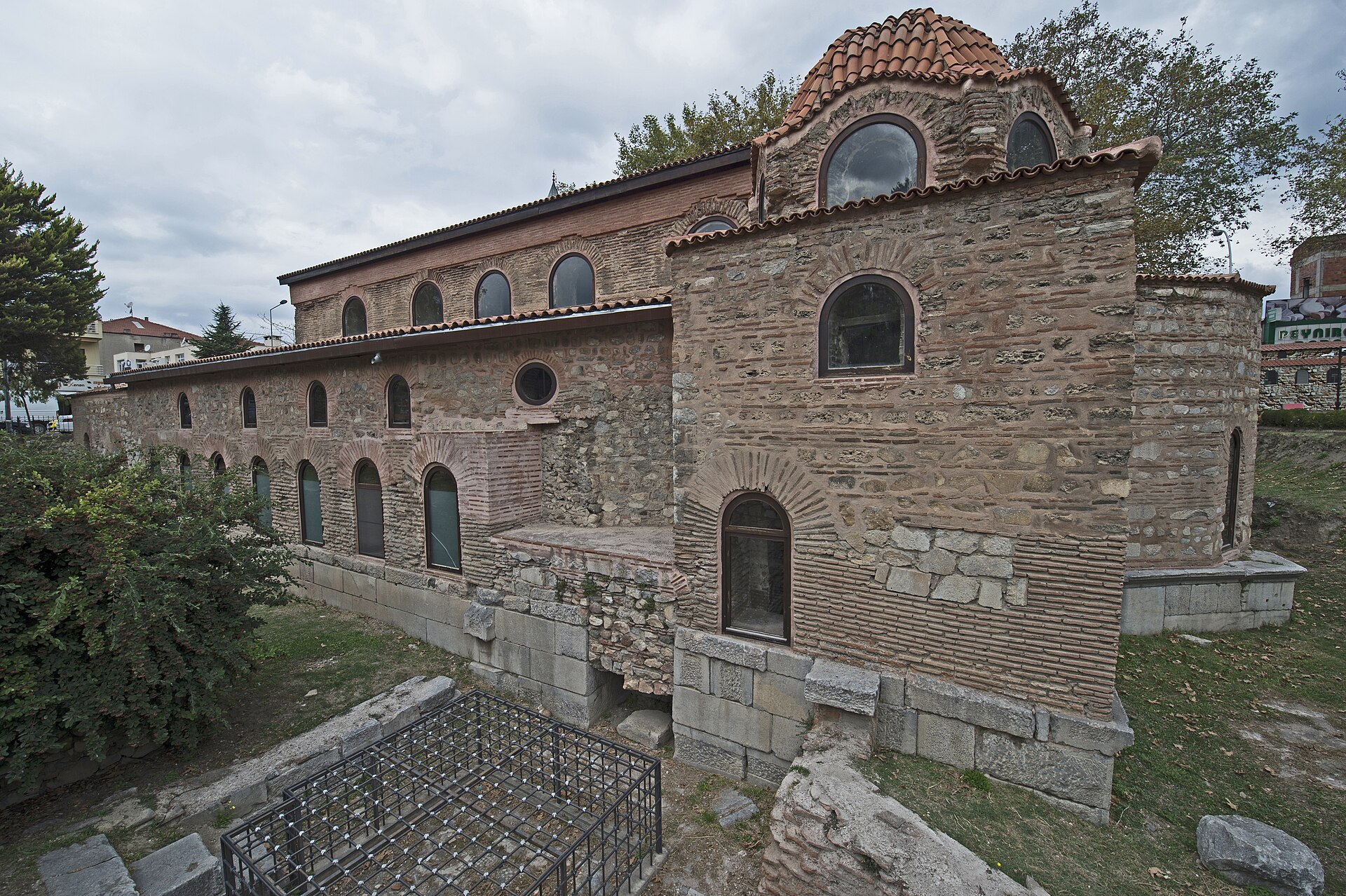 Brick and stone church building with arched windows, a tiled roof and a small dome, standing in a grassy excavated area below street level under a cloudy sky
