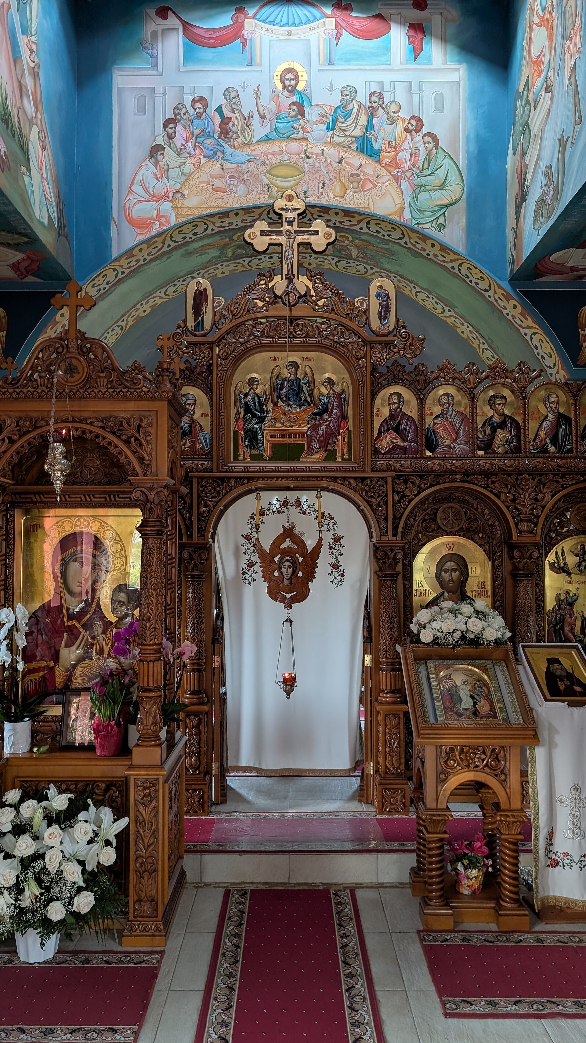 A tall wooden icon screen across the front of a church, with rows of painted icons of saints and Christ on gold backgrounds, a central doorway with carved gates, and lamps hanging in front