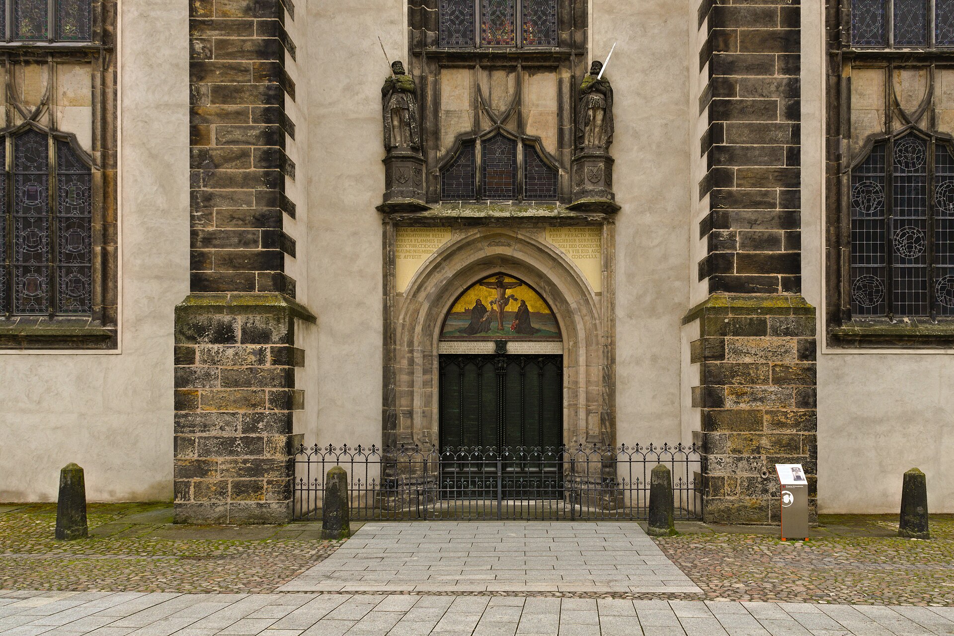 The pointed stone doorway of a Gothic church with tall bronze doors covered in raised Latin lettering, and a painted panel of a crucifixion scene above
