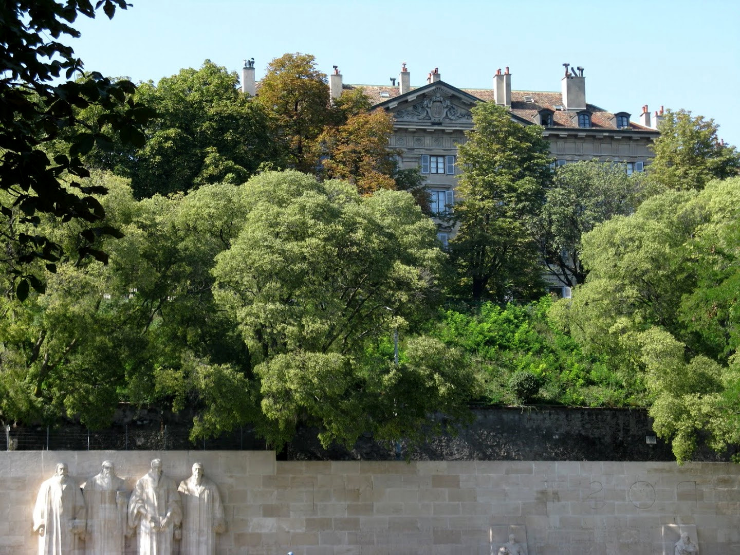 A long stone wall with large standing statues of robed men in the center and carved inscriptions, behind a lawn and trees in a park
