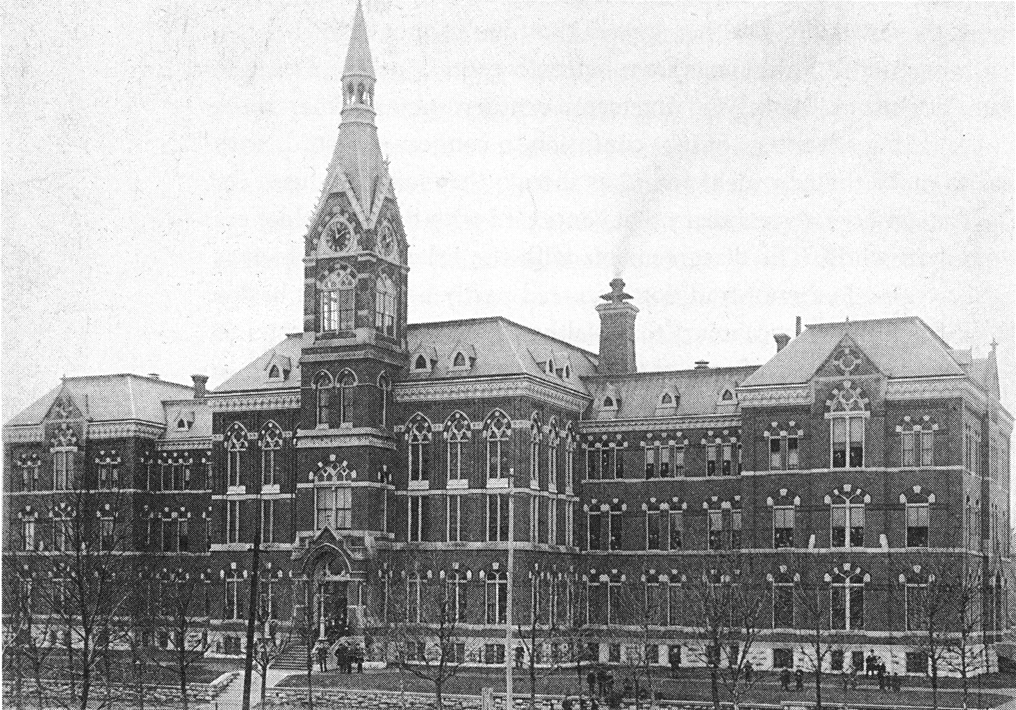 Old photograph of a large brick building with Gothic towers and pointed windows behind a row of young trees