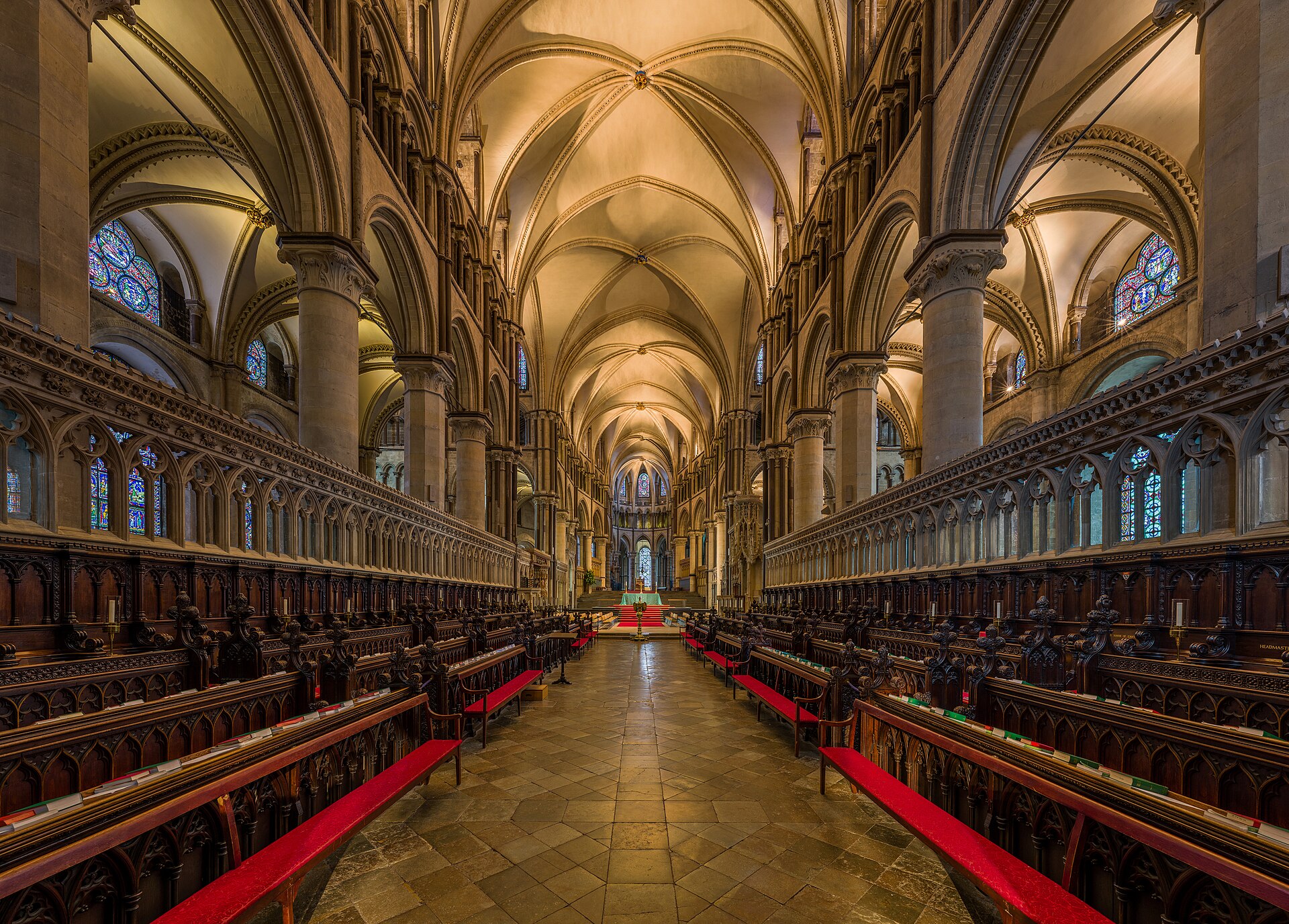 Interior of a Gothic cathedral choir with carved wooden stalls on both sides, a long aisle, pointed stone arches and stained glass windows at the far end