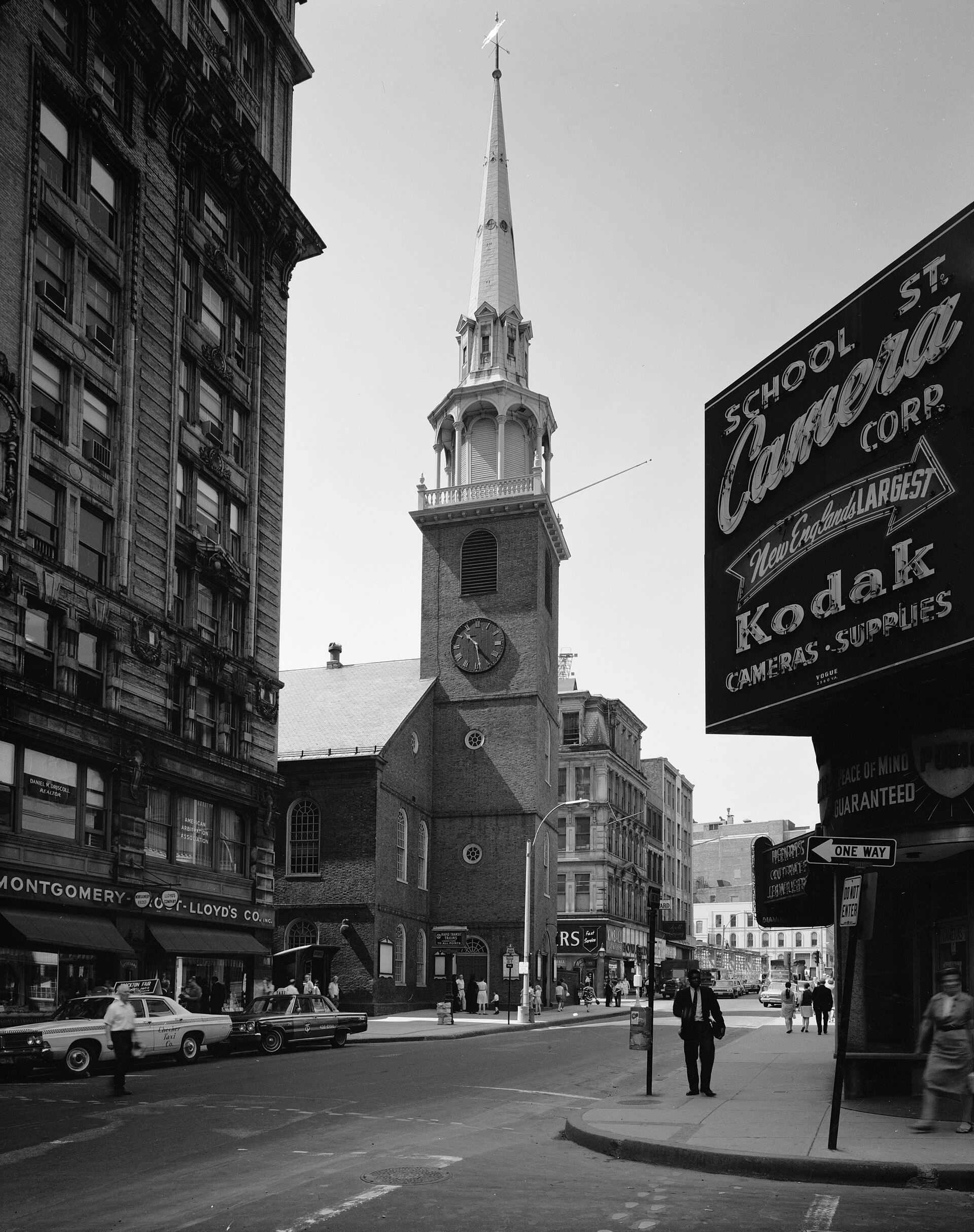 Black and white photograph of a tall brick meeting house with a white wooden steeple rising in stages above a clock, on a city street corner