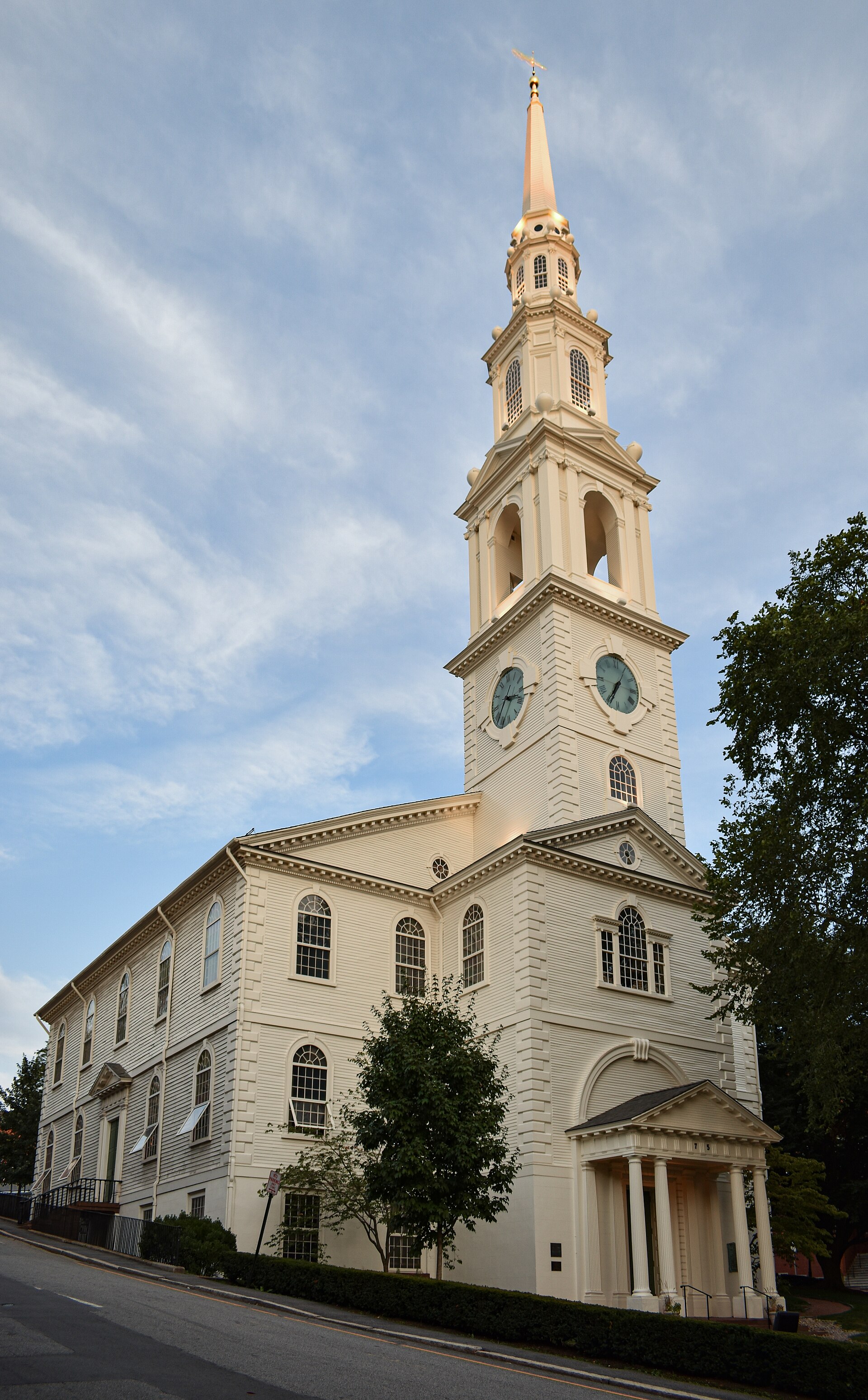 A white wooden meetinghouse with a tall multi-stage steeple, seen from a city street with trees