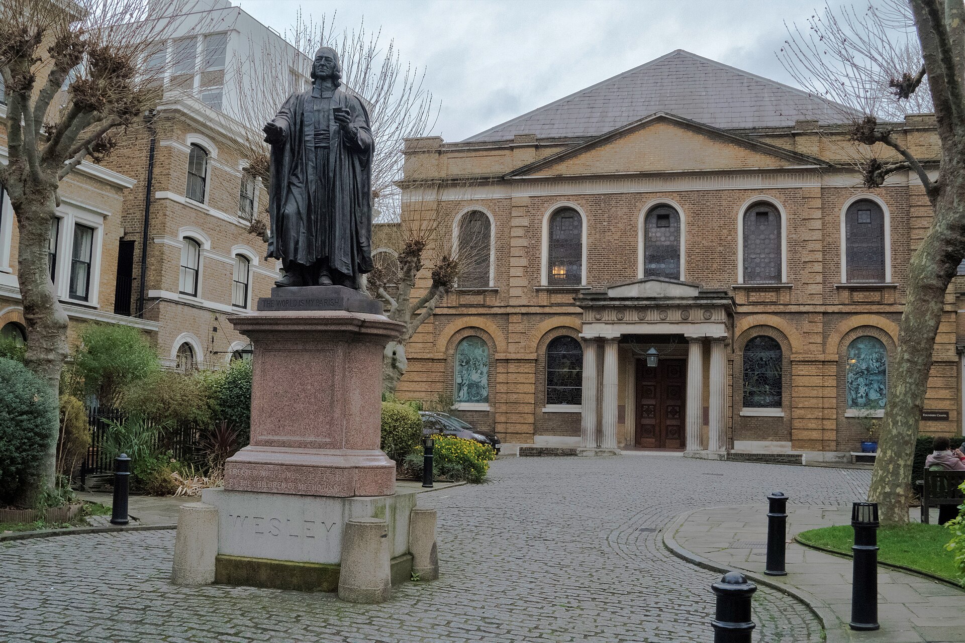 A Georgian brick chapel with a classical pediment and columned porch behind iron railings, with a bronze statue of a preacher in the forecourt