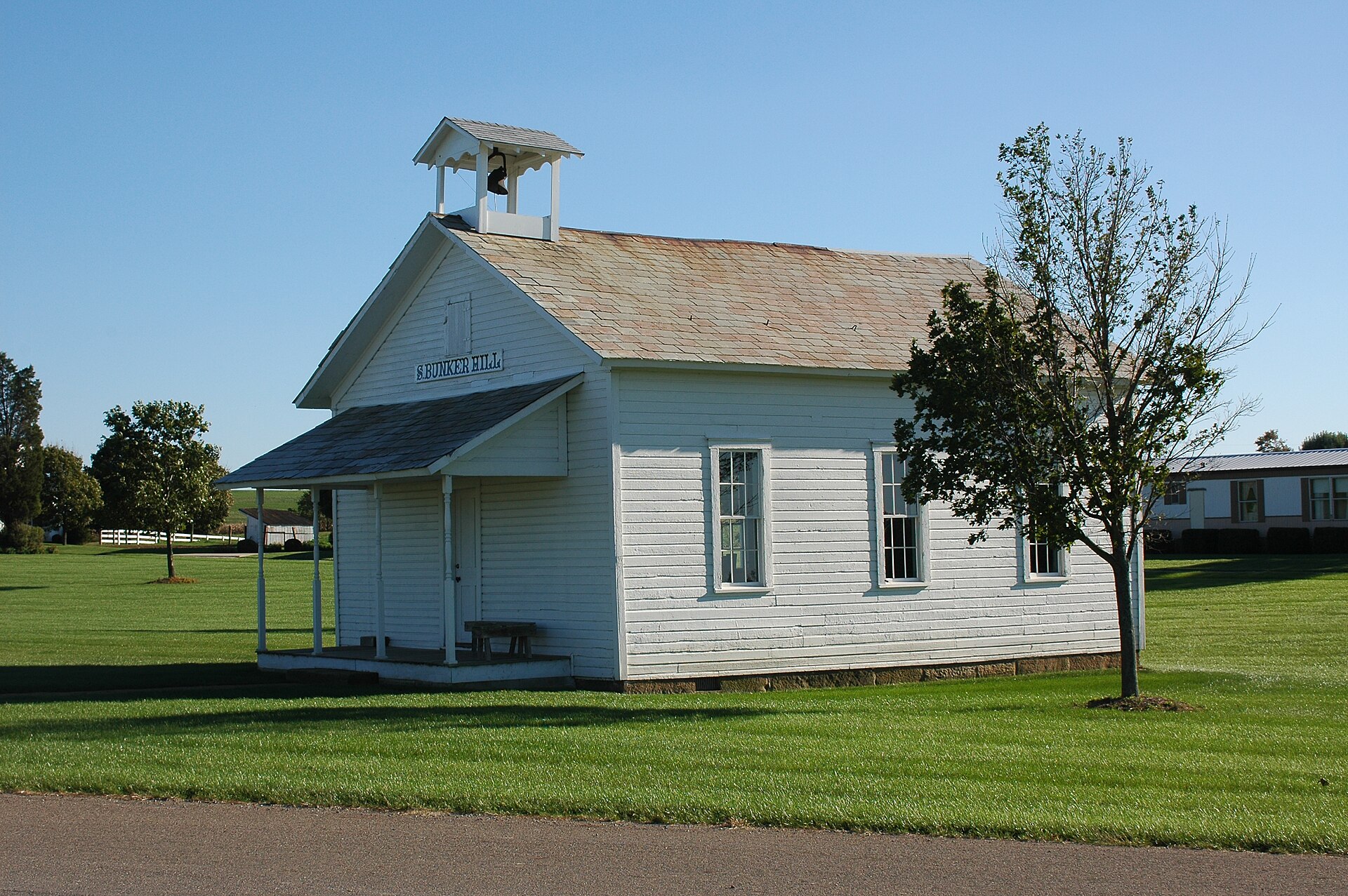 A small white one-room schoolhouse with a bell tower and a short porch standing on a lawn