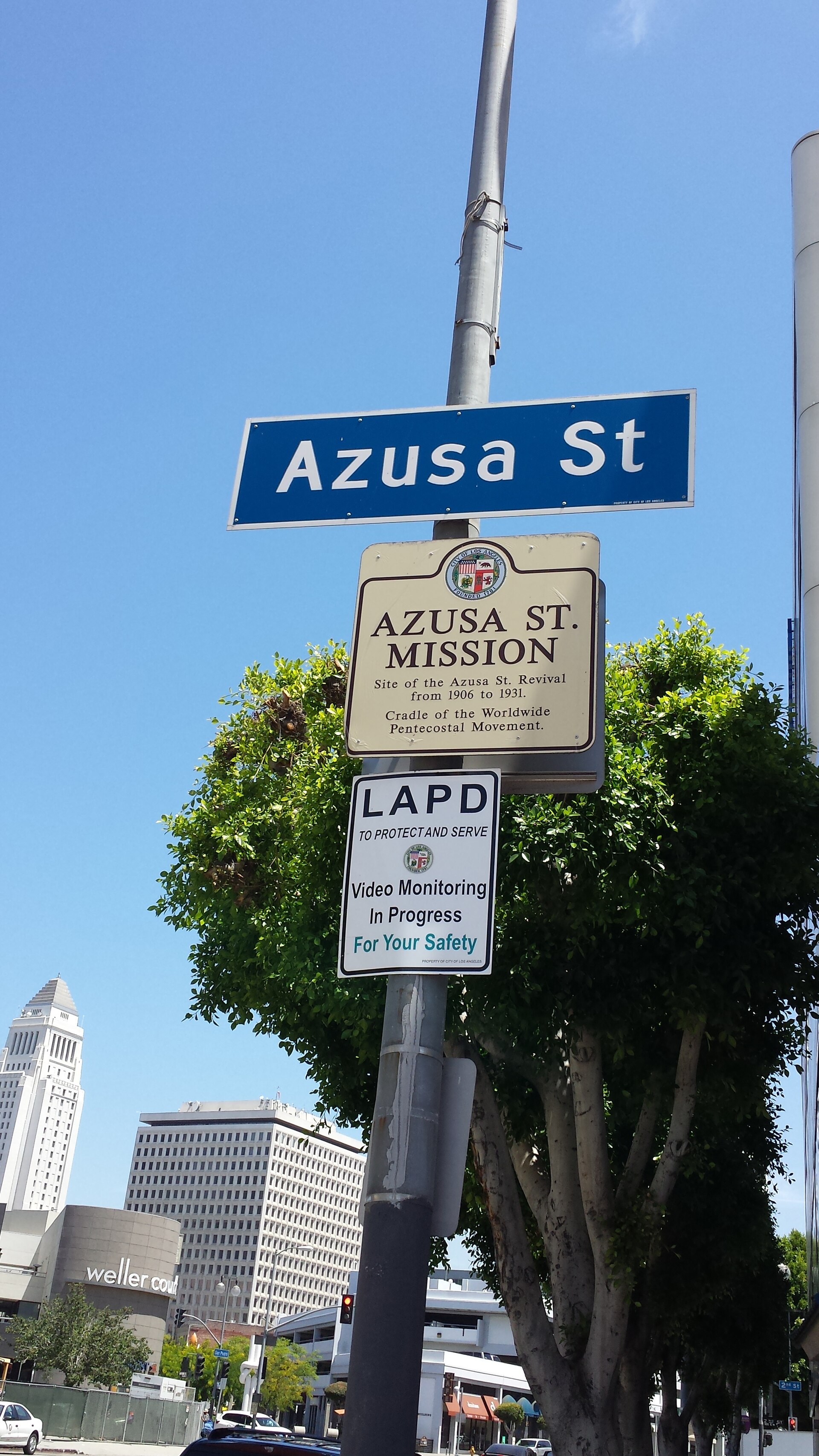 A tall historical marker sign on a city street in Los Angeles describing the Azusa Street revival, with buildings behind it
