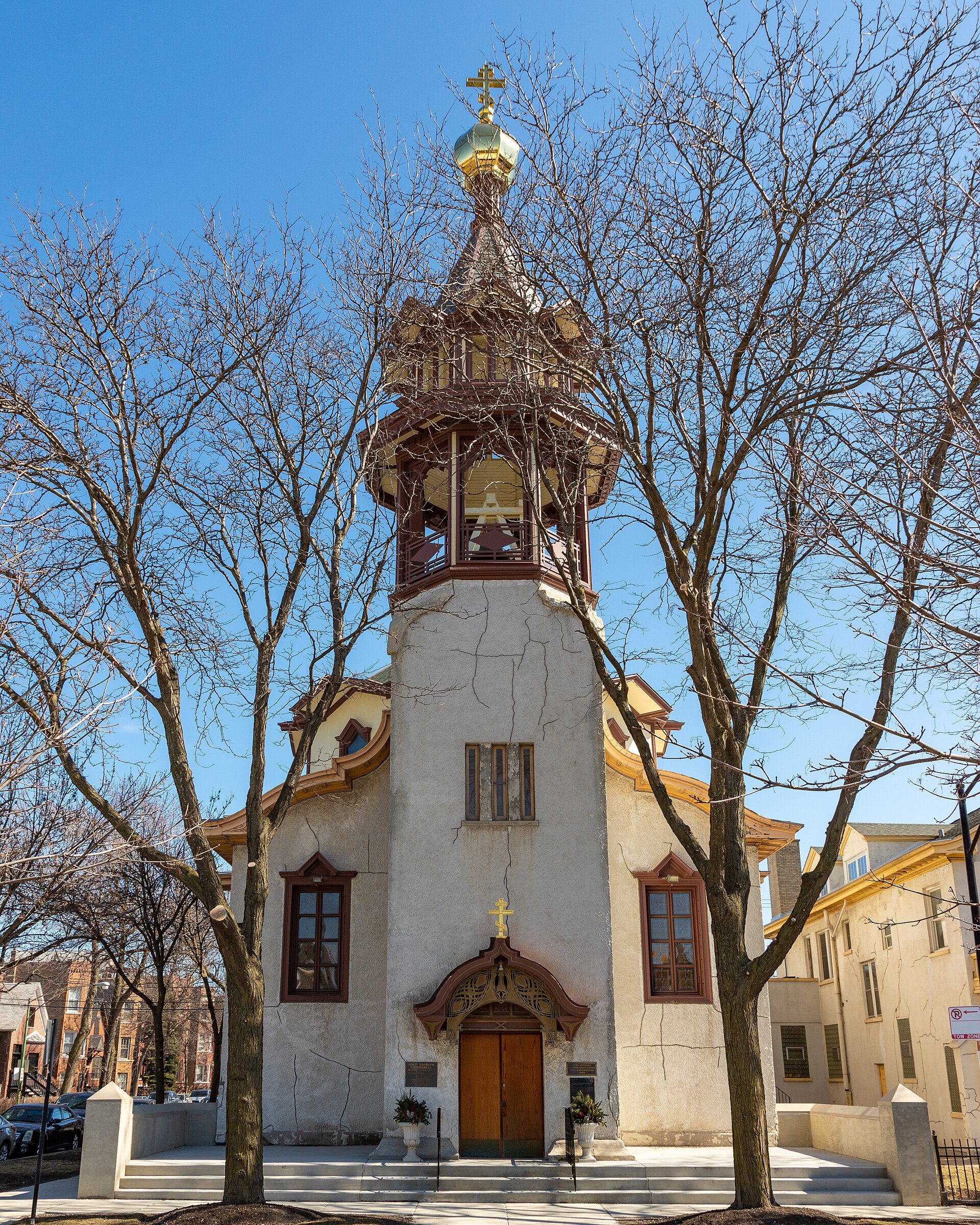 Exterior of a small white and tan Orthodox cathedral with an octagonal bell tower topped by a dark onion dome and a cross, on a city street