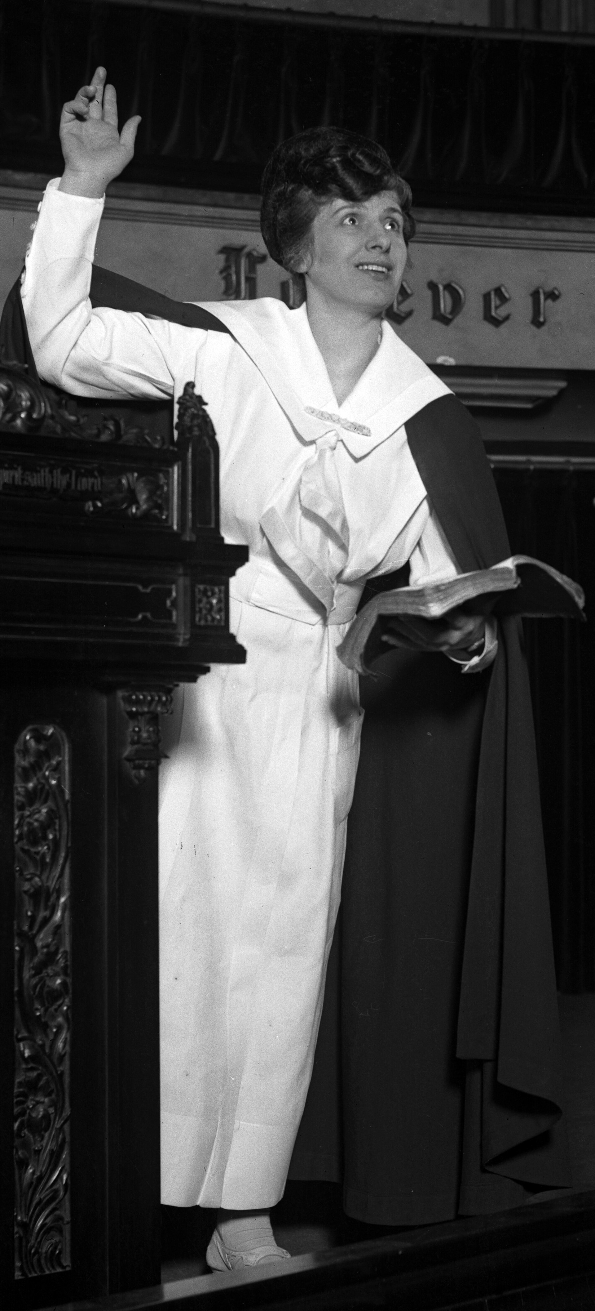 Black and white photograph of Aimee Semple McPherson in a white dress, arms raised, preaching from a platform in front of a large audience inside a domed auditorium
