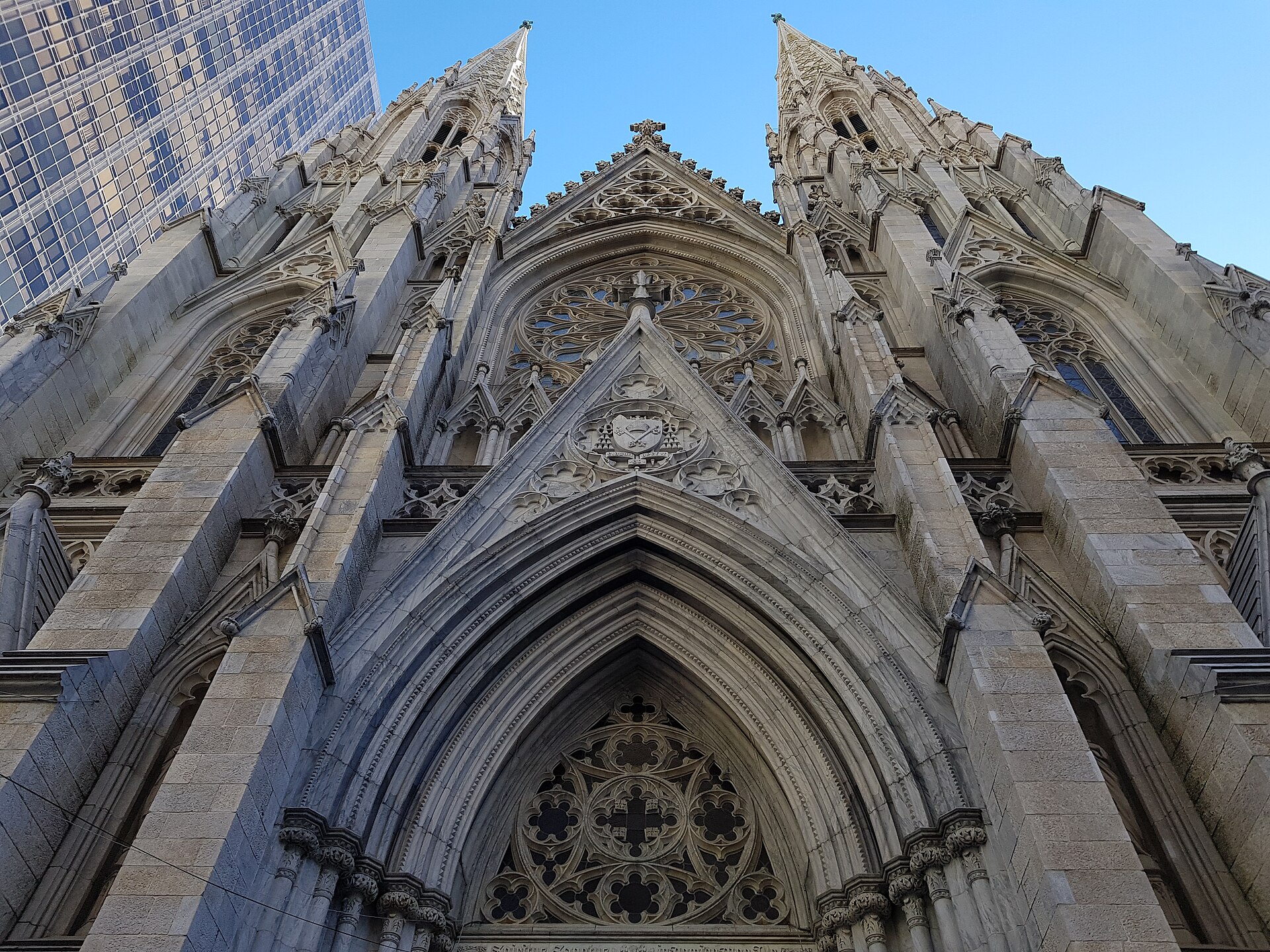 Front of a large white Gothic Revival cathedral with two tall spires and a rose window above three pointed doorways, between Manhattan buildings
