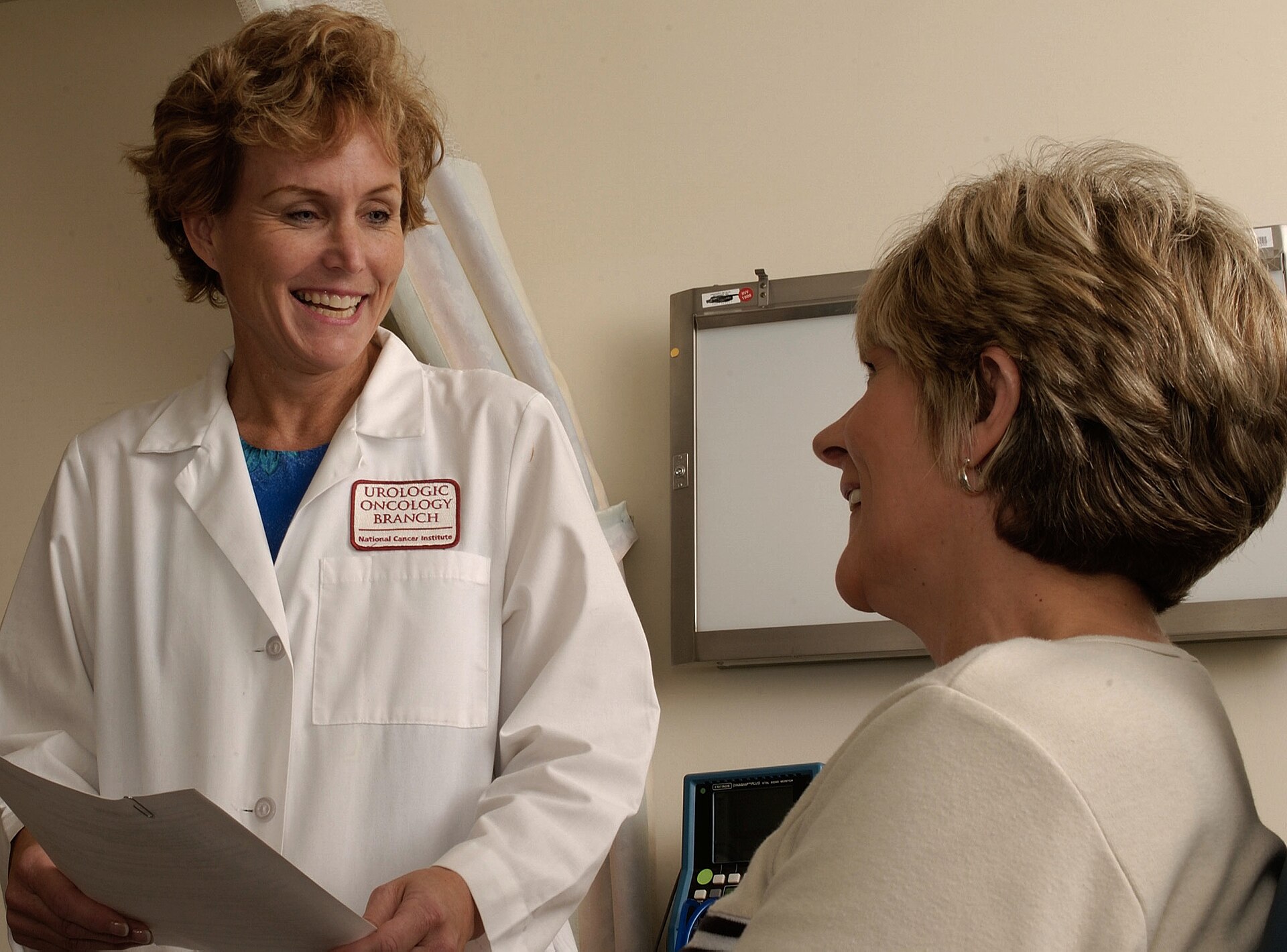 A doctor in a white coat sits facing a patient and talks with her in an exam room
