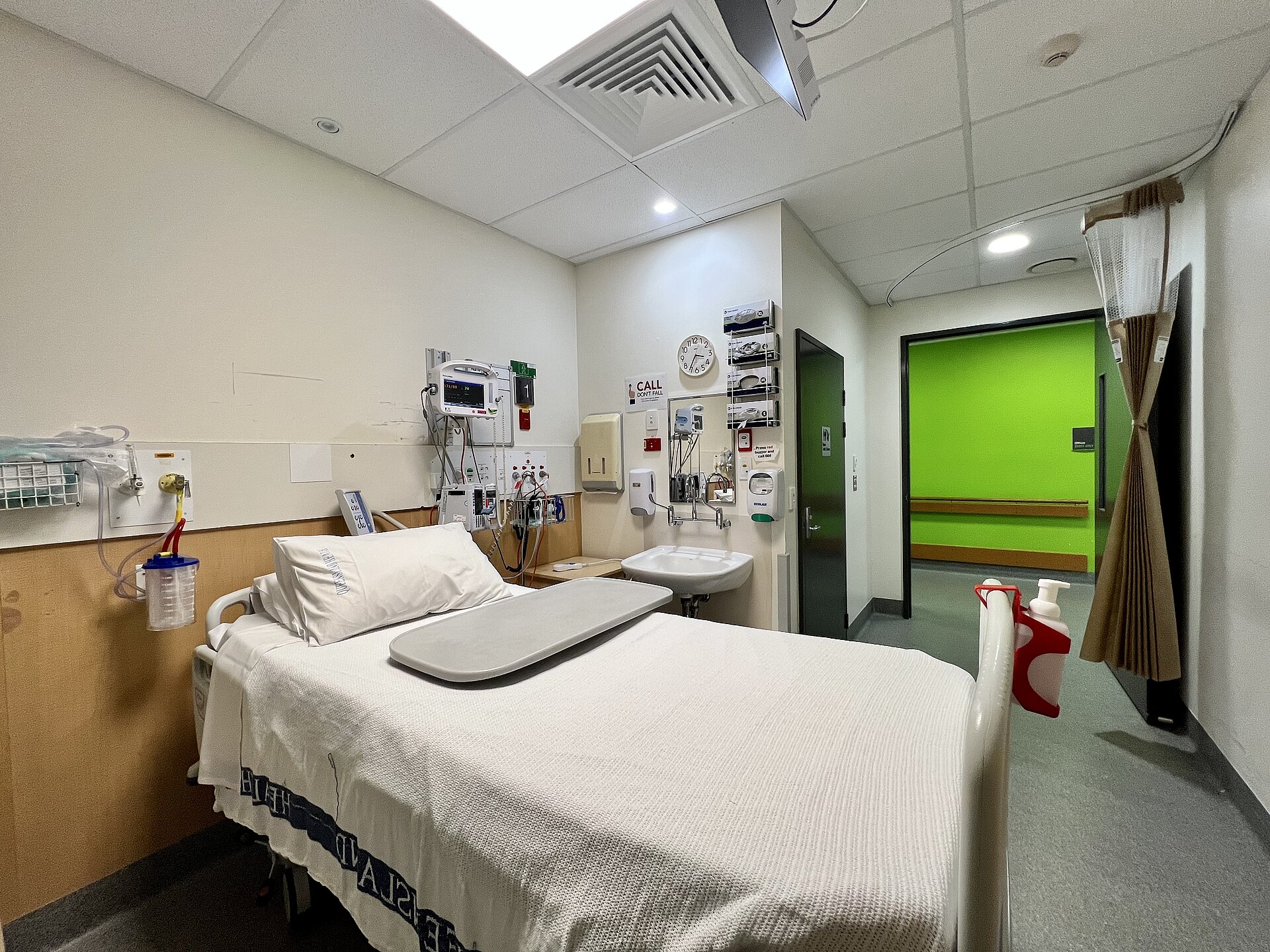 A single-bed hospital room with a bed, a chair, overhead lights, wall equipment and a window