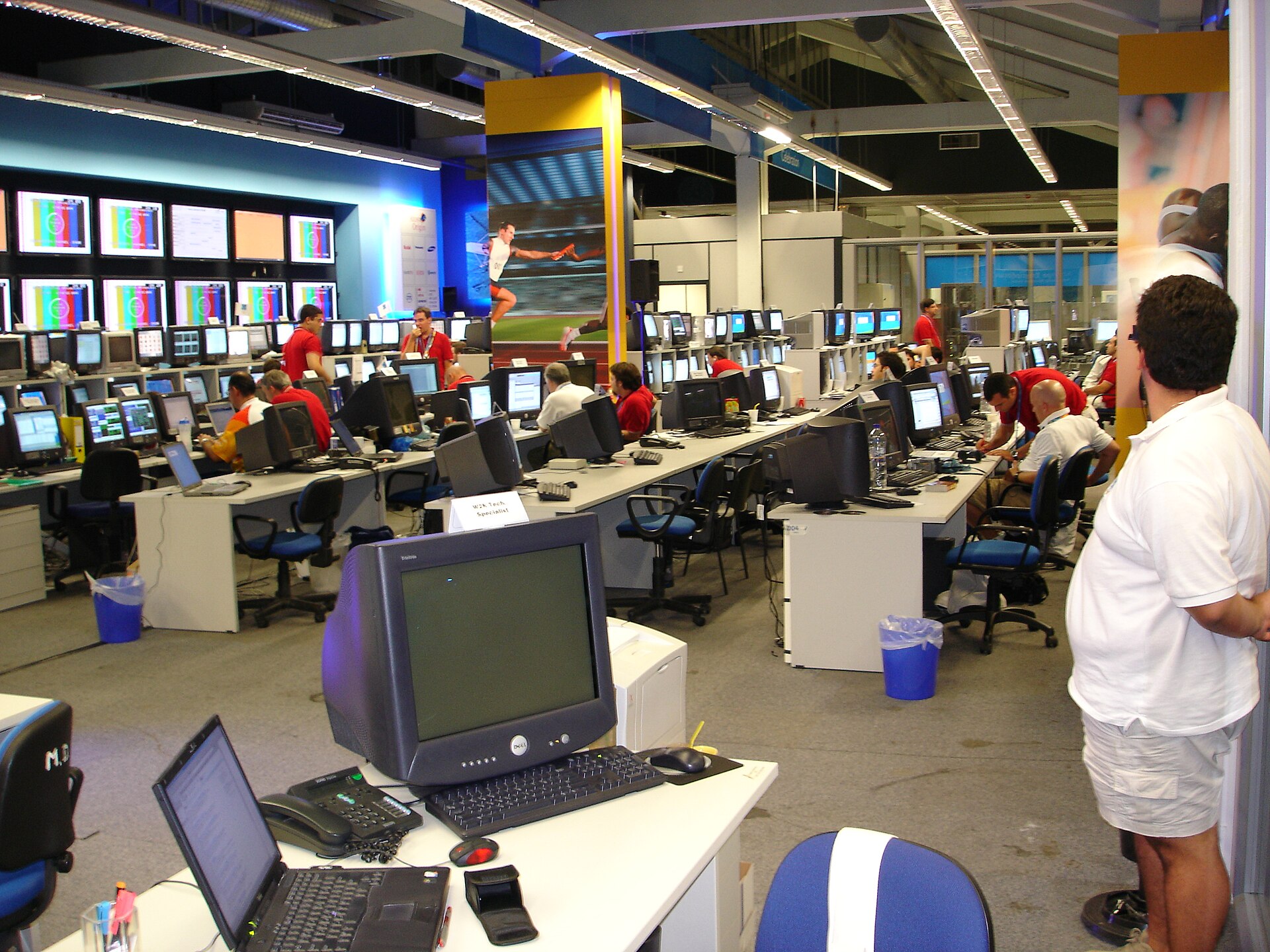 A large room of workers at rows of desks with computer monitors facing a wall of large display screens in a technology operations center