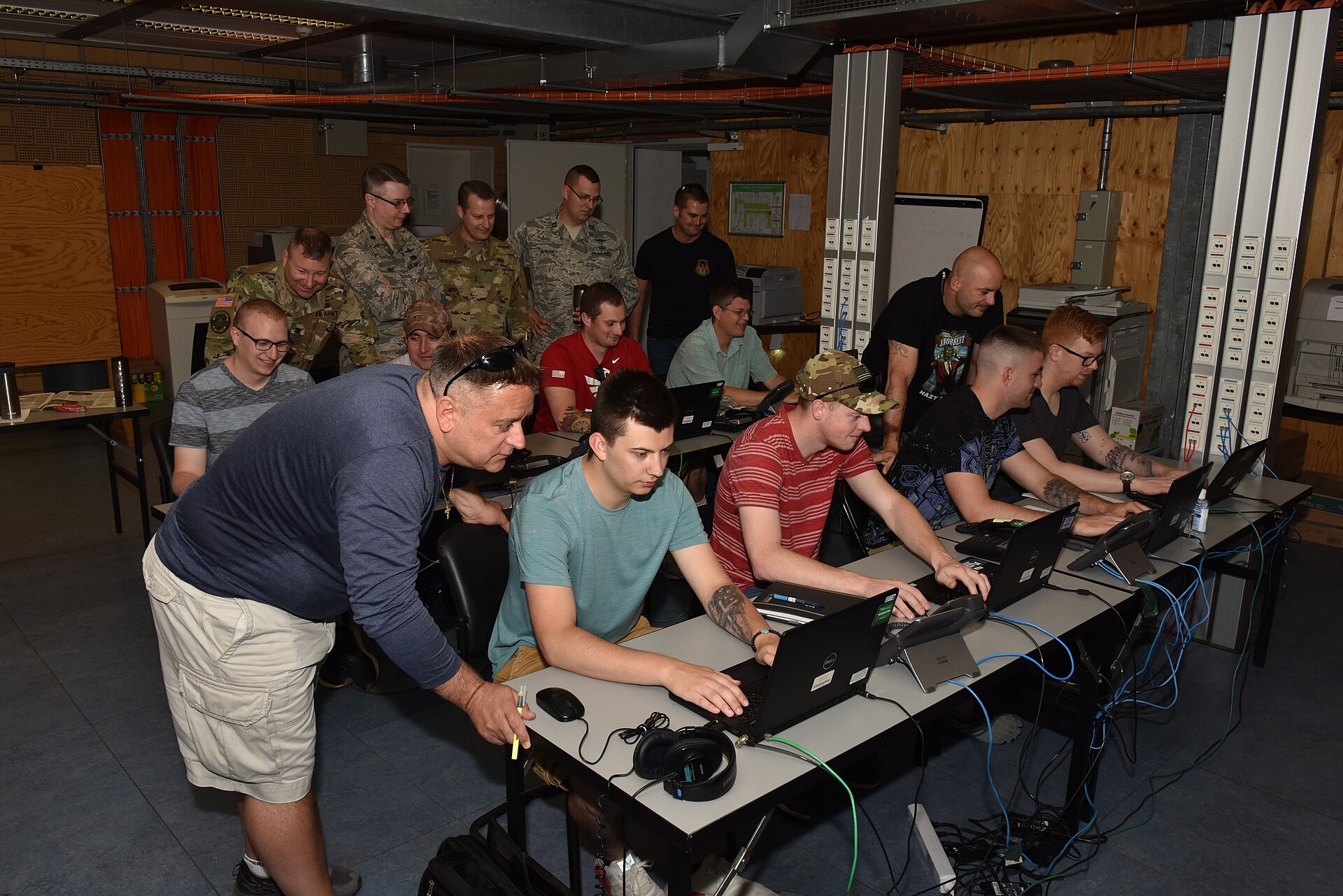 Several uniformed service members sit at a long row of desks with laptops and monitors, working side by side in a training room