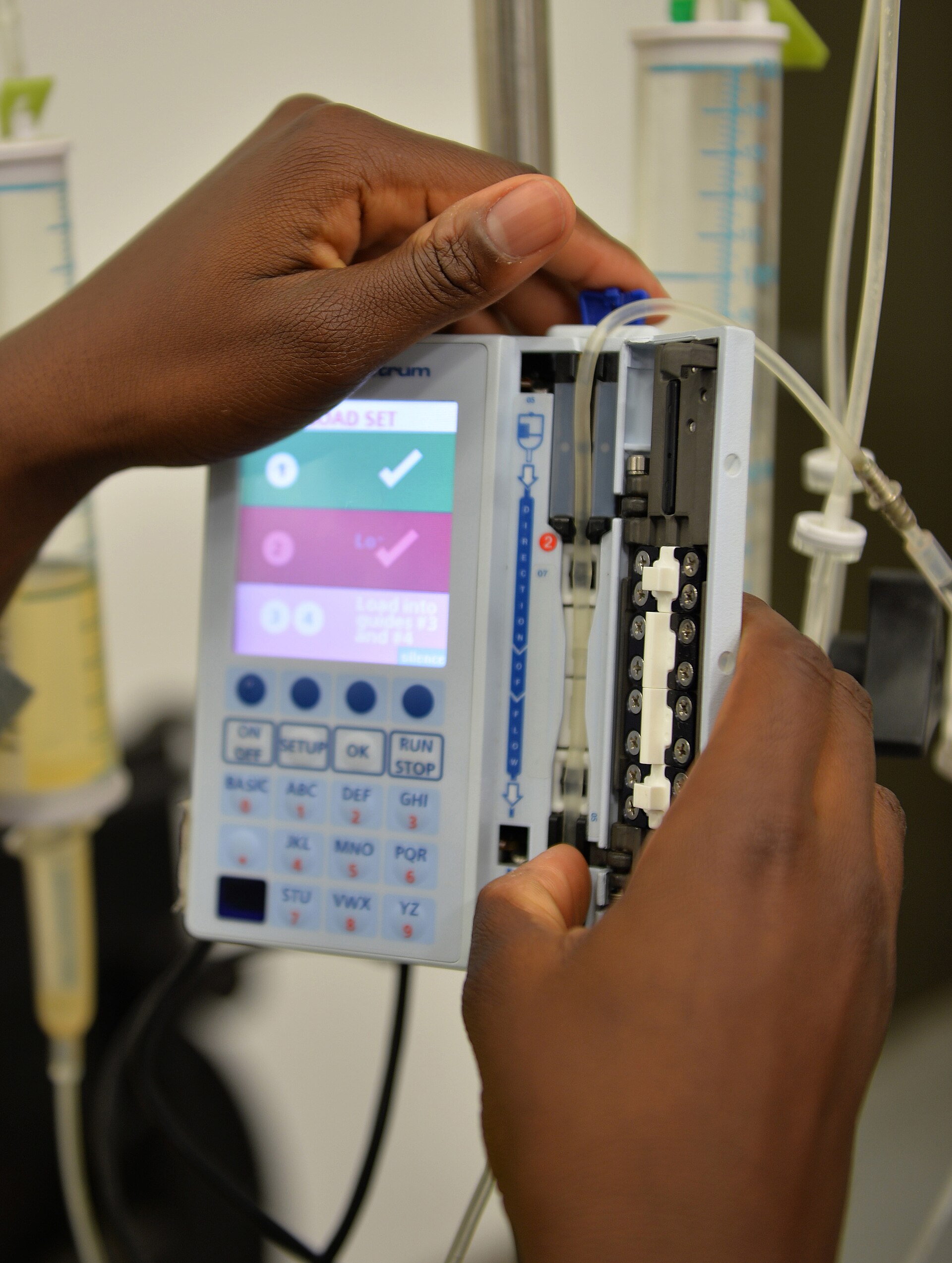 A uniformed technician in a workshop presses buttons on the front of a tall infusion pump mounted on a pole, with test equipment on the bench behind him