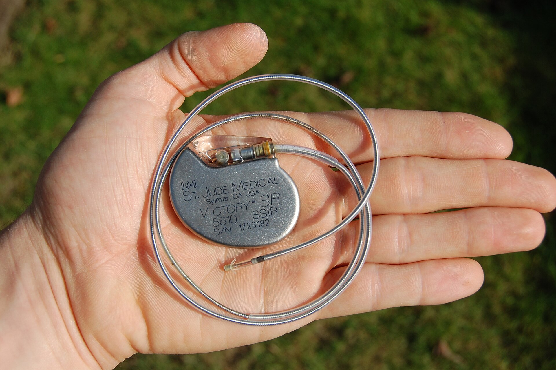 A hand holding a small, rounded metal pacemaker case with a long coiled lead wire attached