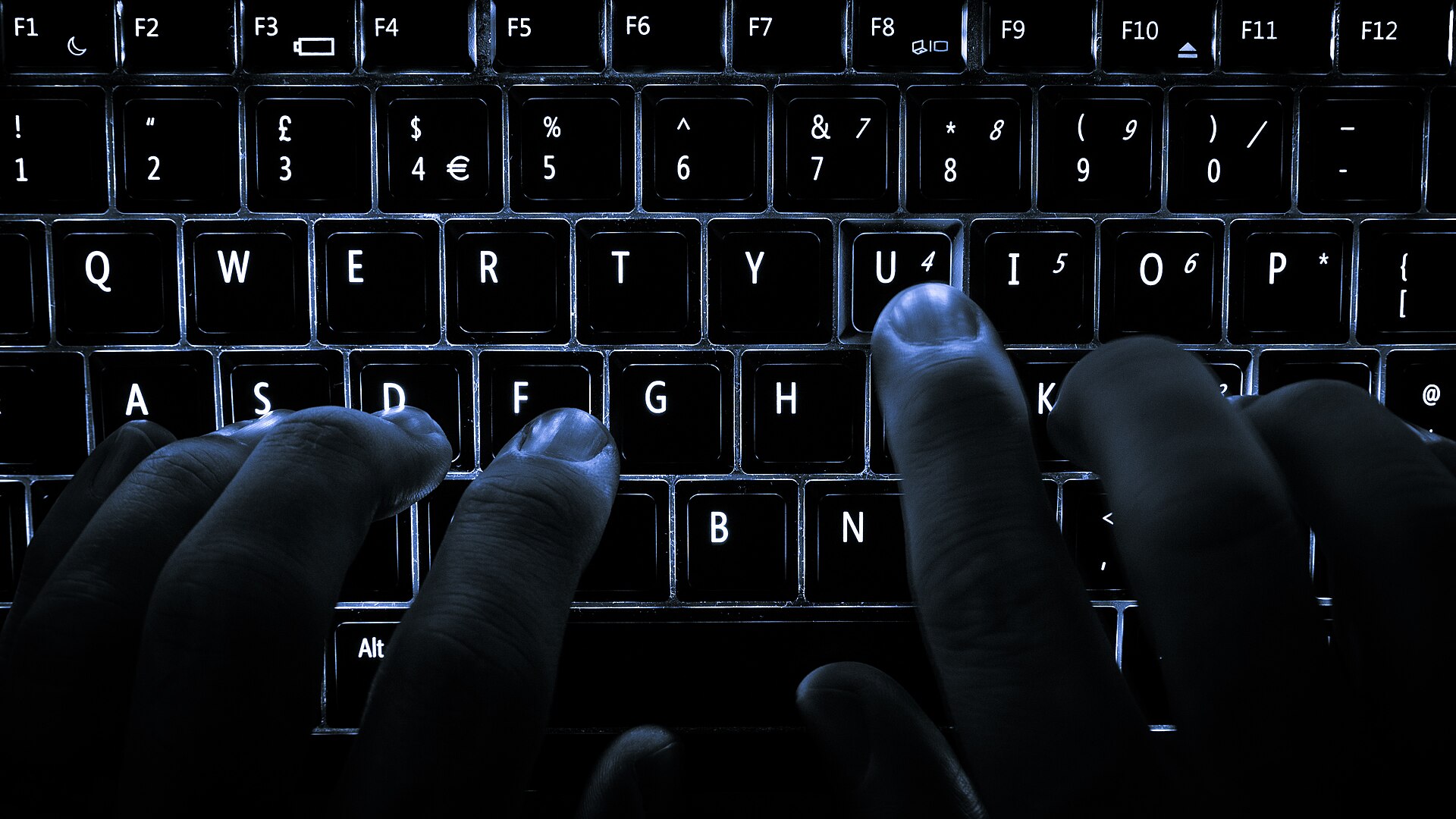 Close-up of a backlit laptop keyboard in a dark room, with a person's fingers resting on the home row keys and one finger pressing the U key.