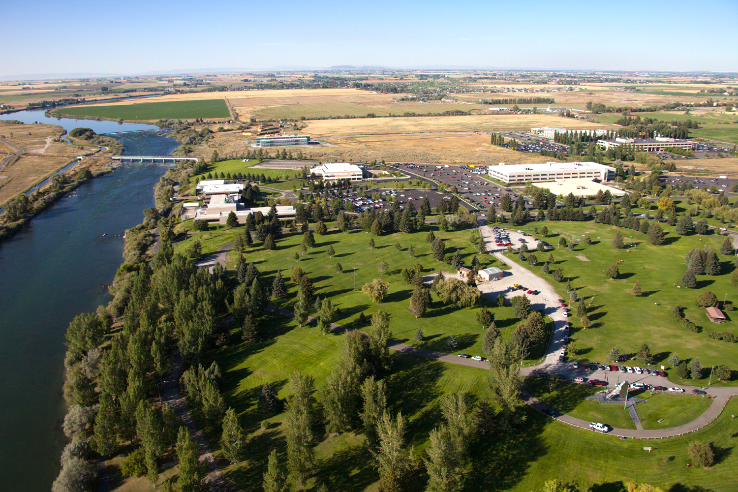 Aerial view of low office and laboratory buildings with large parking lots, set among lawns and evergreen trees beside a wide river crossed by a bridge, with farm fields stretching to the horizon.