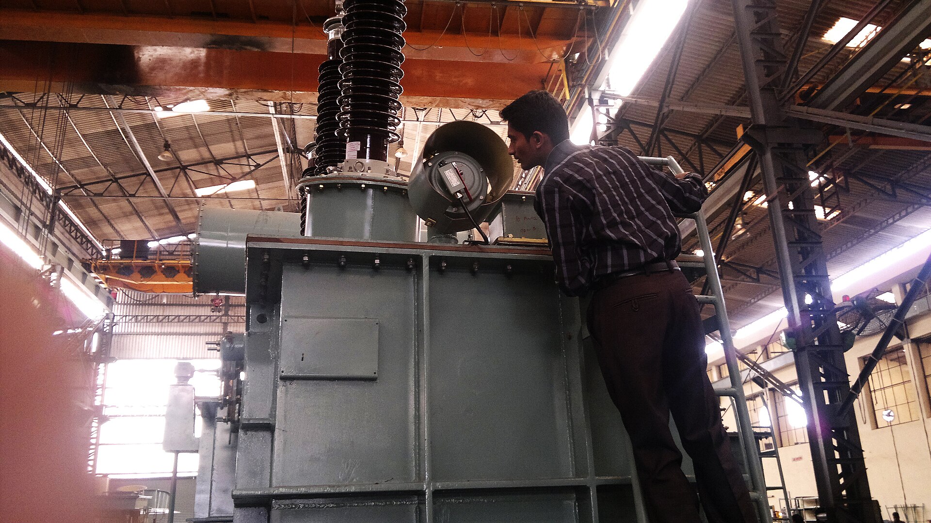 An engineer in a hard hat stands beside a large outdoor oil-filled power transformer and inspects a pressure relief valve mounted on top of it.