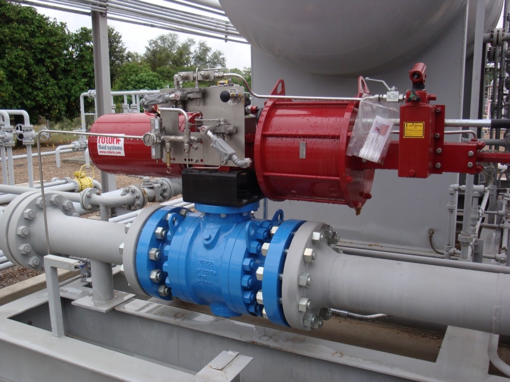 An industrial ball valve in a pipeline with a large pneumatic actuator mounted on top, fitted with tubing, a control box and a hand-operated hydraulic override.