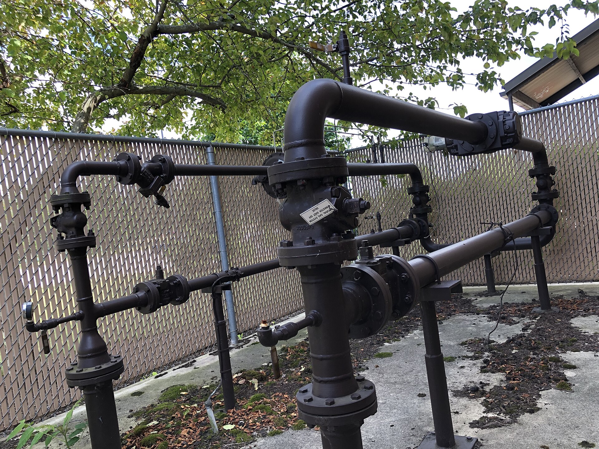 Above-ground natural gas pipeline equipment: painted pipes, valves and fittings rising from the ground inside a fenced enclosure on a grassy campus.