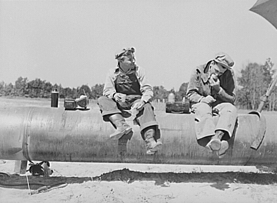 Black and white photo of a work crew in hats standing along a large steel pipeline laid in a trench